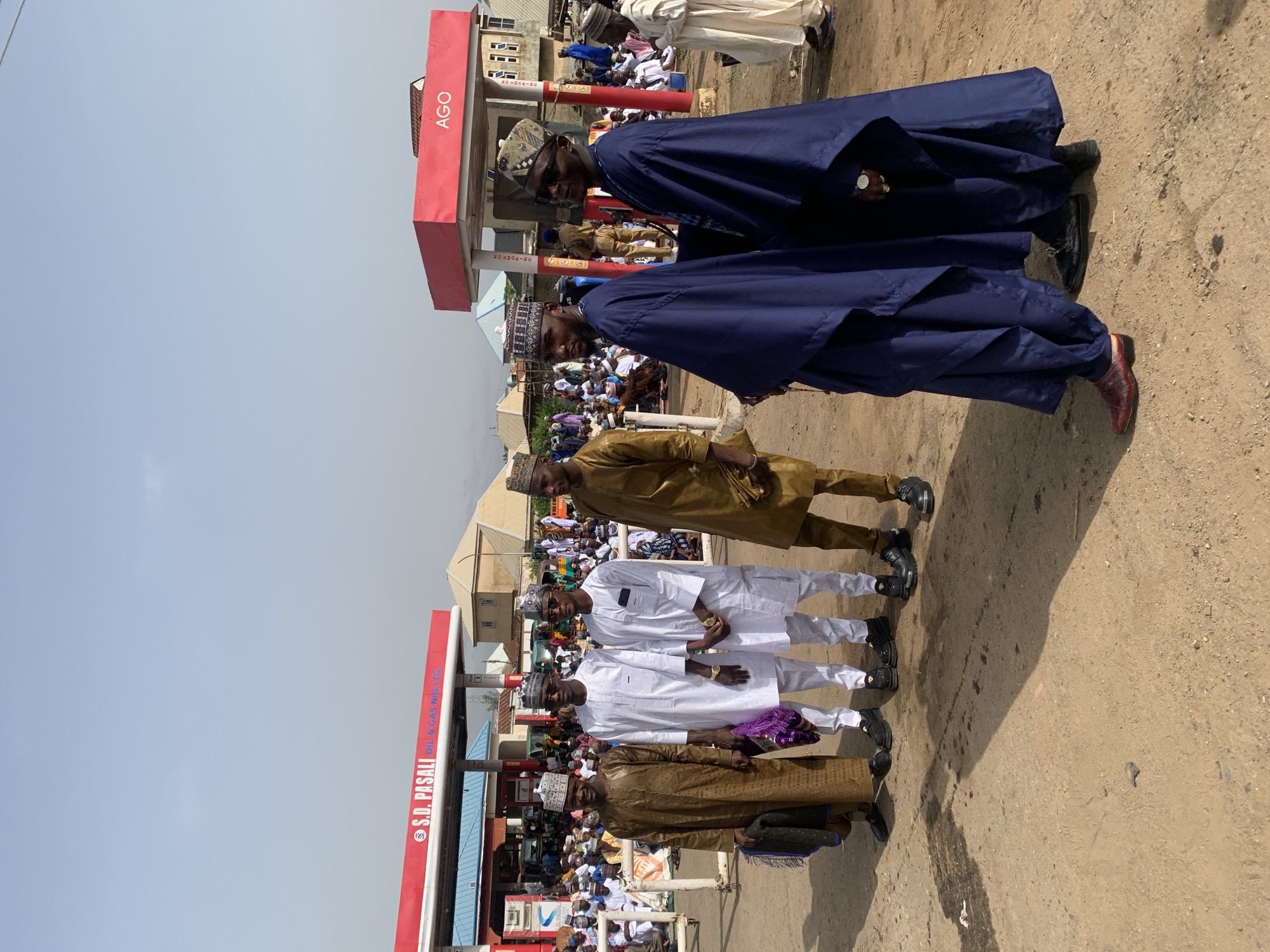 The image depicts a group of men standing outside a gas station in what appears to be Jos, Nigeria. The gas station has the name "AGO" and "S.D. Pasali Oil & Gas Nig Ltd" visible on the red canopies. The men are dressed in traditional Nigerian attire. The group seems to be posing for a picture. Some are wearing long blue robes, others are in white, and some in brown outfits. Several men wear traditional caps. A crowd is gathered in the background. The ground is a mix of dirt and pavement. The sky is partly cloudy.