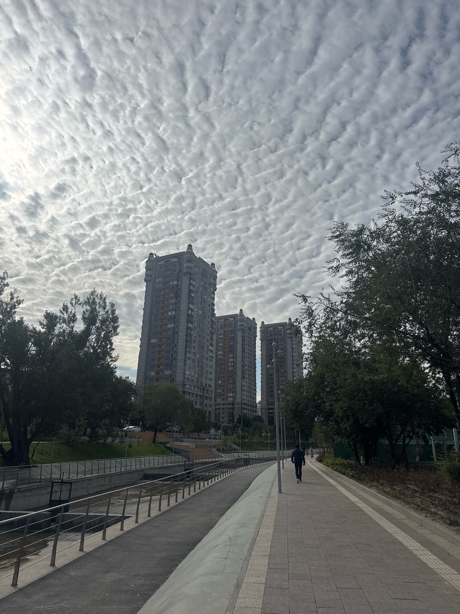 The image captures a wide-angle view of a modern urban environment under a dramatic sky filled with altocumulus undulatus clouds. In the mid-ground, three tall, grey residential buildings with distinct architectural features, including decorative elements at their peaks, dominate the skyline. To the left of the buildings, a tree-lined canal or dry riverbed runs parallel to a paved pedestrian walkway. A metal railing lines the edge of the walkway. A lone individual, seen from behind wearing dark clothing, walks away from the camera on the right side of the paved path. The foreground shows the textured surface of the walkway, composed of grey and lighter grey rectangular paving stones, bordered by a textured tactile paving strip. Greenery, including trees and grass, flanks the walkway and the buildings. In the far distance, a white car is visible on a road that ascends a slight incline behind the canal. The overall scene suggests a well-maintained public space within a city. This scene is identified as being located in the city of Almaty, Kazakhstan.