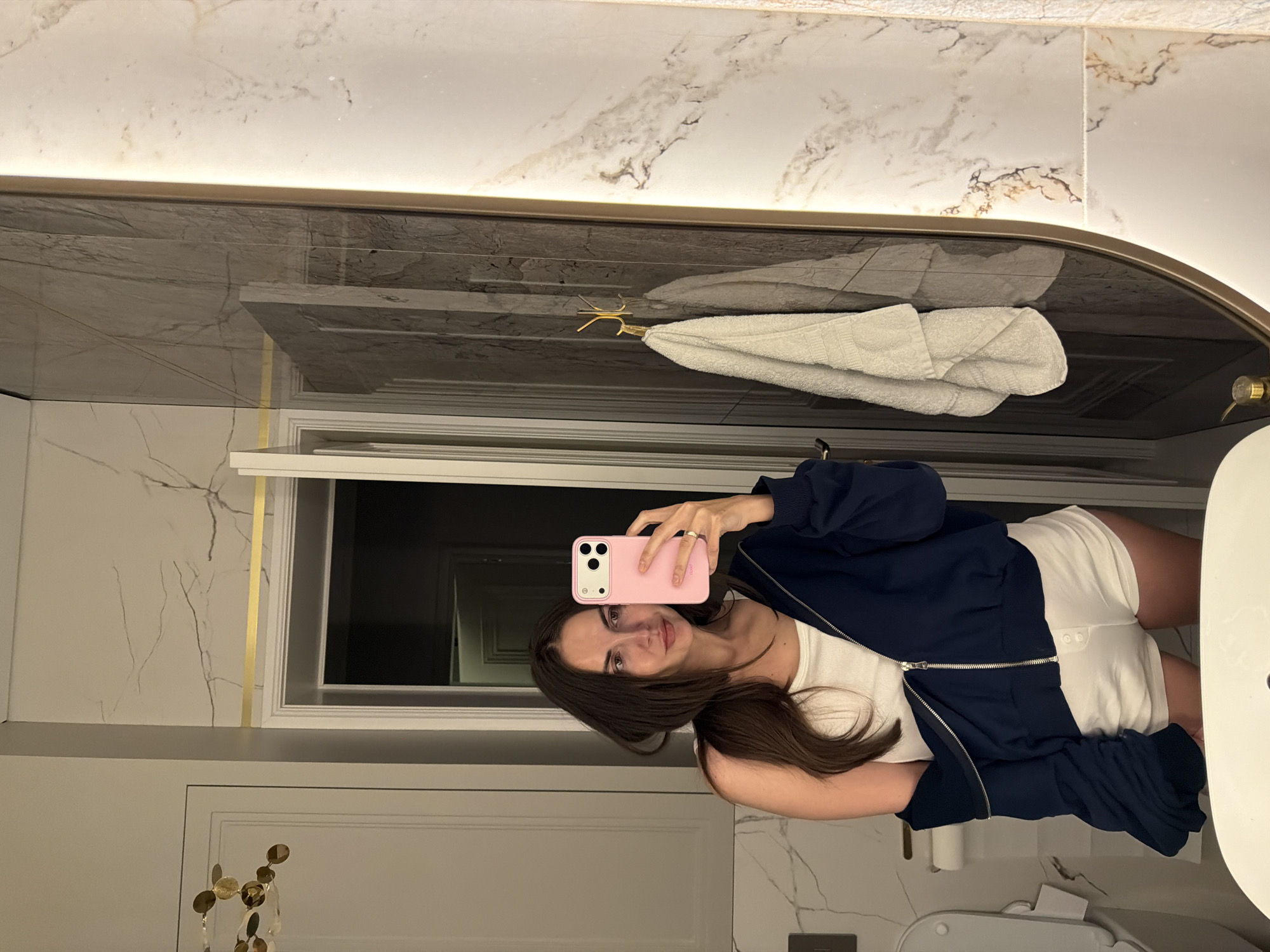 A woman stands in a bathroom, taking a selfie in the mirror with a pink smartphone. She has long brown hair and is wearing a white top, white shorts, and a navy zip-up jacket. The bathroom features white and gray marble walls, a gray marble countertop, a white towel hanging from a gold hook, a white door with gold decorations, and a white toilet and sink. The location is Krasnoyarsk, Russia.