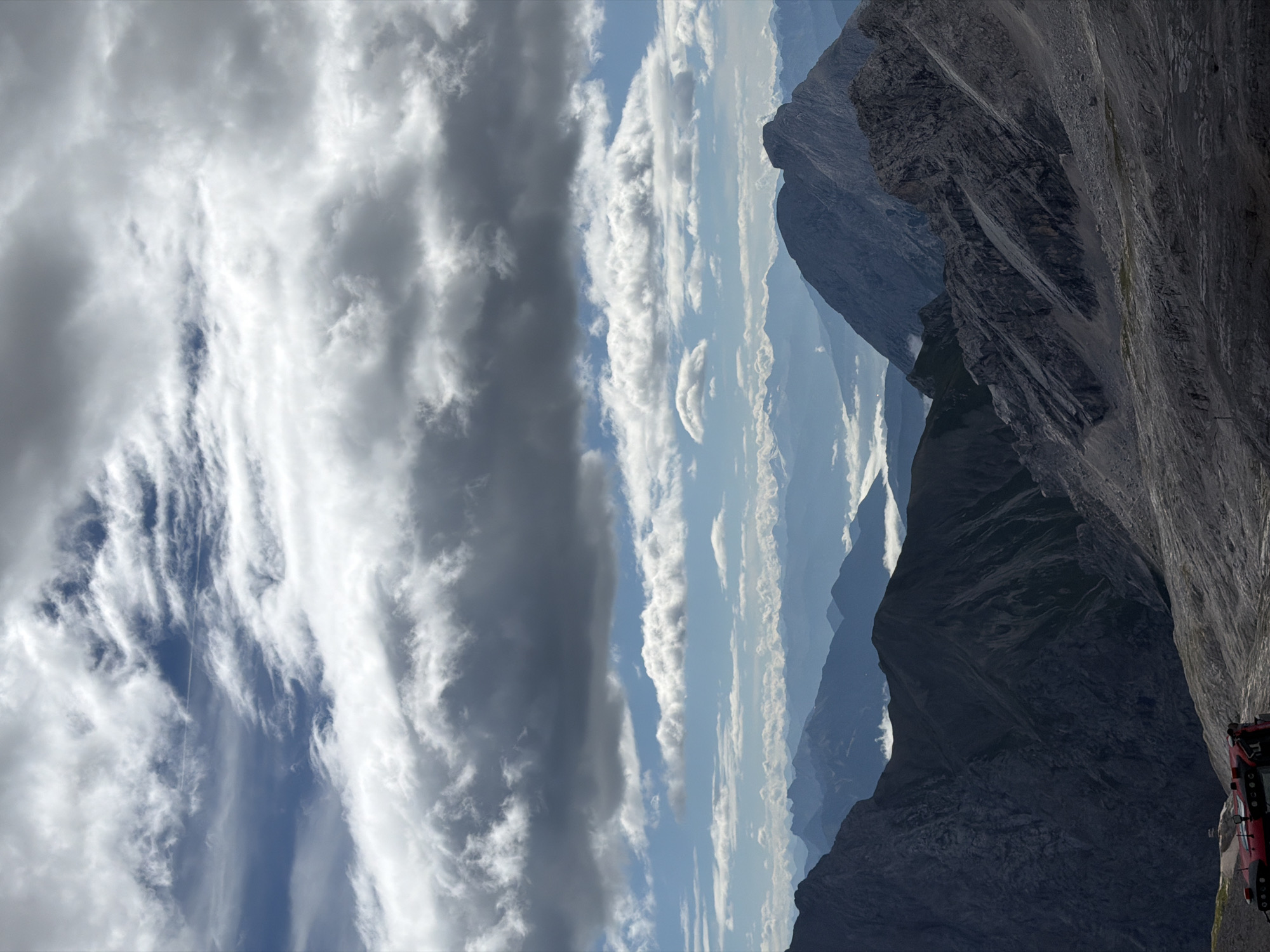 The image captures a high-altitude mountain landscape, likely the Zugspitze, the highest peak in Germany, near Garmisch-Partenkirchen. The foreground is dominated by rugged, rocky terrain with a red vehicle, possibly a snowcat or utility vehicle, visible on the lower left. In the mid-ground, dark, imposing mountain ranges recede into the distance, their slopes carved by geological formations. A layer of white clouds hovers in the valleys between these peaks, creating a sense of depth and atmosphere. The background reveals a vast expanse of layered mountains fading into a hazy blue, with a clear sky punctuated by dramatic, white and gray clouds. The overall scene evokes a sense of grandeur and the raw beauty of alpine environments.