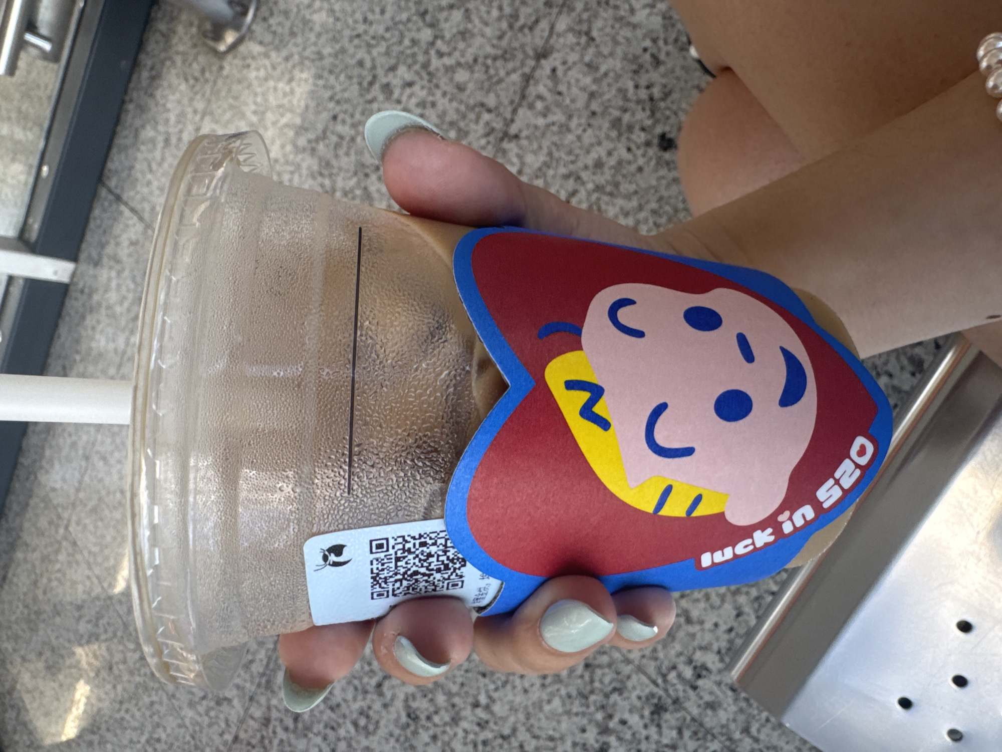 A person is holding a plastic cup with a sticker in their right hand. The sticker has a heart shape with a face on it and the words "luck in 520". The cup has a lid and contains a brown liquid. The person's fingernails are painted a light green color. The scene appears to be set outdoors on a gray tiled surface, possibly in Nan'an District, China. The image is taken from a high angle.