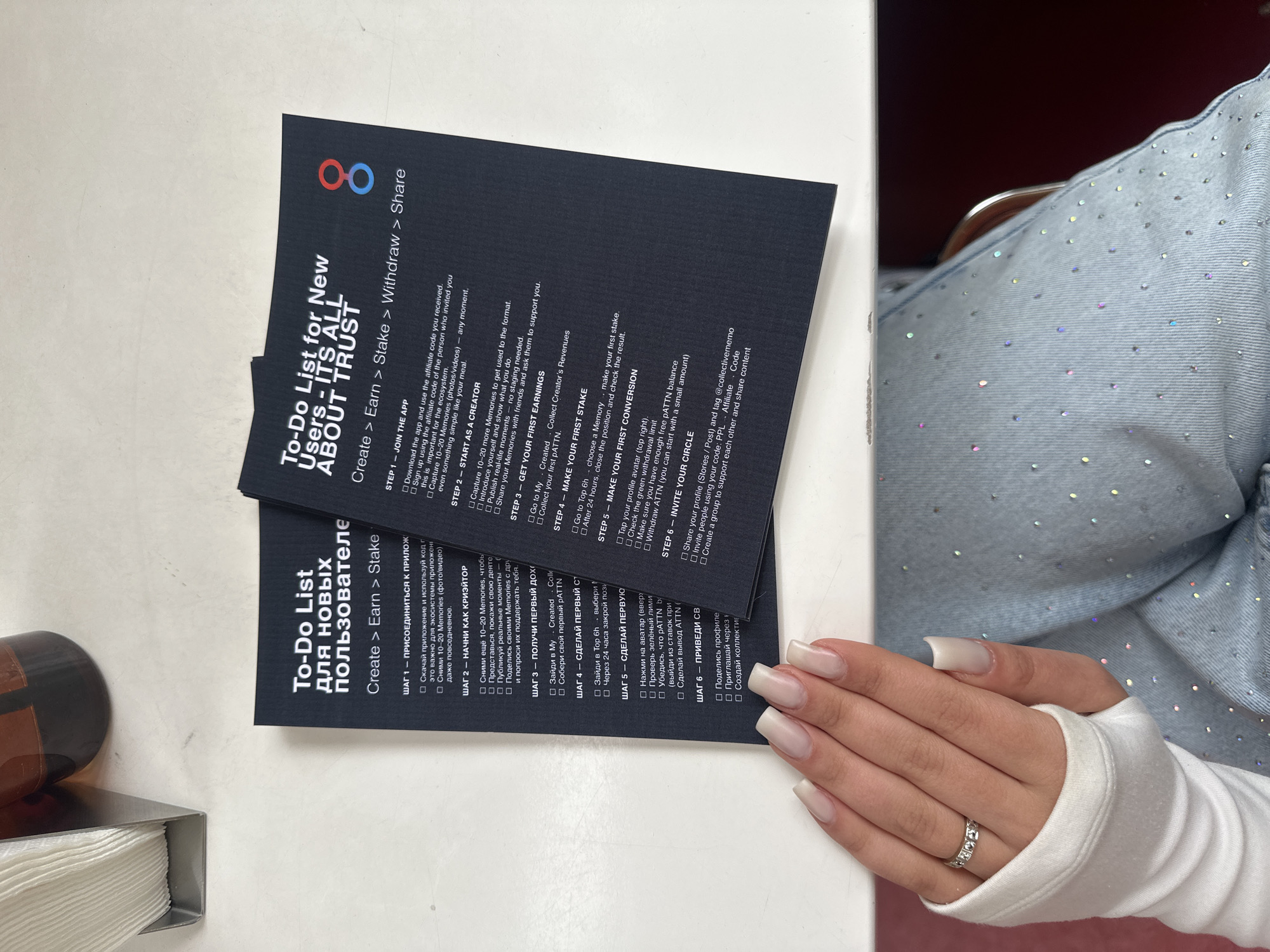Here is a detailed description of the image:
**Scene:** The image depicts a close-up view of two "To-Do List" cards for new users of an application, one in Russian and one in English, placed on a white surface, likely a table. A portion of a person's lap, adorned with bedazzled light blue denim jeans and a white long-sleeved shirt, is visible to the right. The person's hand with manicured nails and a ring is resting near the cards. In the lower left corner, there is a glimpse of what seems to be a napkin dispenser or holder. The focus is on the cards and the hand.
**Details:**
* **Cards:** Both cards share a similar layout, with the application's logo at the top (a stylized figure eight in red and blue). The cards list steps for new users, such as joining the app, starting as a creator, earning money, staking, conversion, and inviting others. One card is in English, and the other is a Russian translation.
* **Person:** The person's hand is noticeable due to the well-manicured nails and the ring worn on one of the fingers. The clothes suggest a casual or fashionable style.
* **Location:** The presence of a napkin holder hints at the scene being in a café or restaurant.
**City of Yerevan, Armenia:** There is no specific information in the image to suggest that this scene is happening in Yerevan, Armenia.