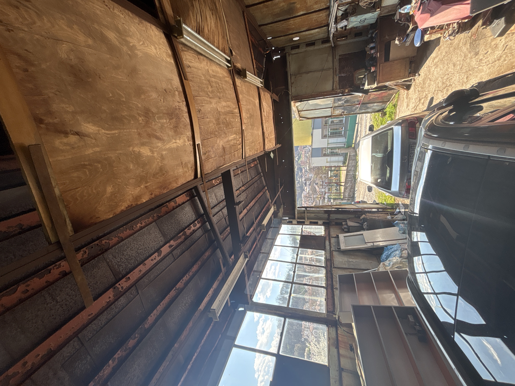 The image depicts the interior of a dilapidated, likely abandoned structure in Zlatograd, Bulgaria. The walls are composed of a combination of plywood panels and weathered metal sheets with visible rust. A series of old windows provide a view of the cityscape, and the sky is visible through broken panes. Various discarded objects, including what appears to be shelving units and other debris, clutter the space. Outside the structure, a silver car is parked, partially visible in the foreground. In the background, a view of Zlatograd is seen through the opening, showcasing buildings and greenery. No people are visible.