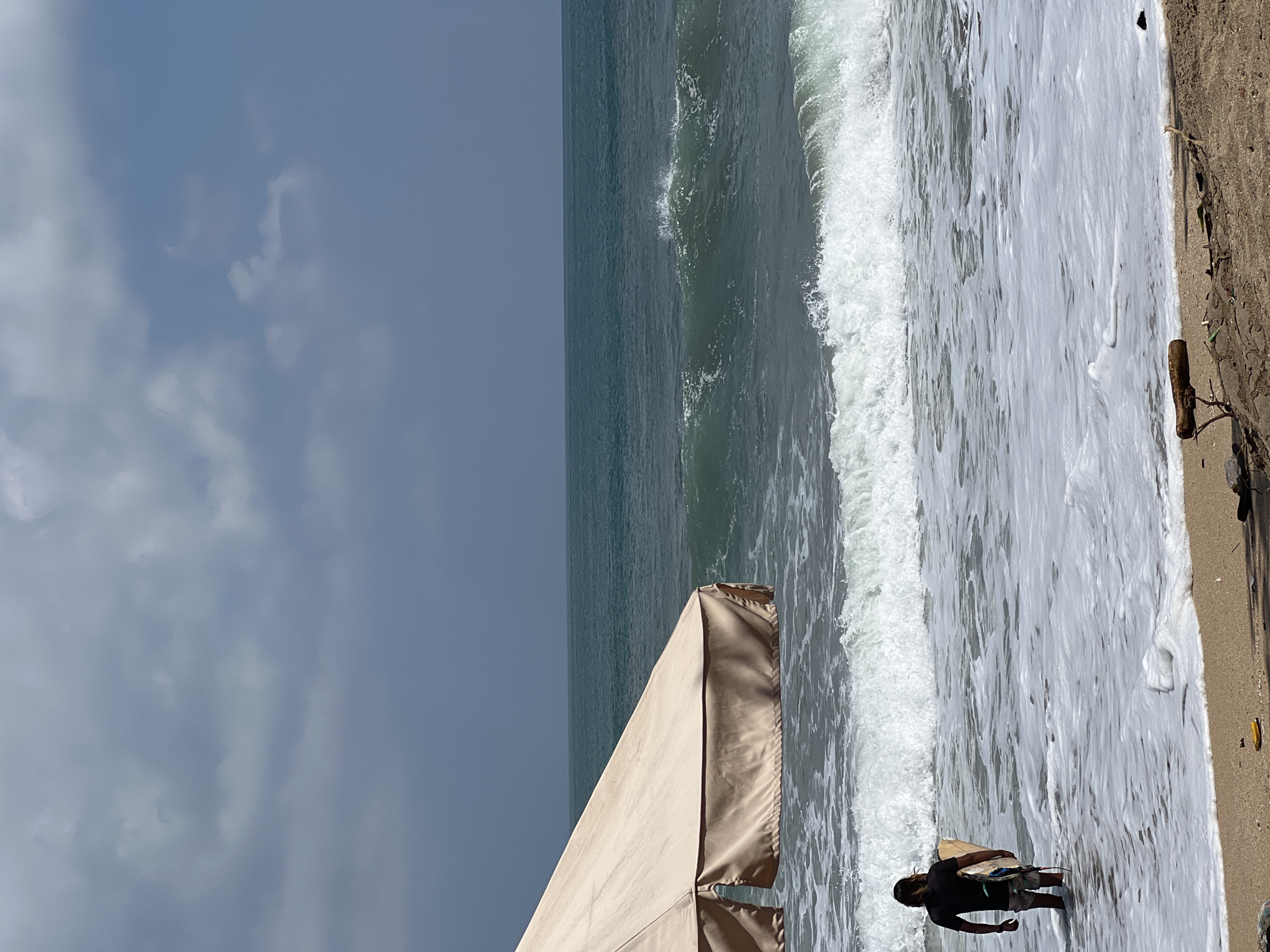 A lone surfer stands in knee-deep water at Kuta Beach, Indonesia, holding a surfboard. The surfer, wearing a black t-shirt and patterned shorts, faces away from the camera, looking towards the ocean. A large, beige beach umbrella is positioned to the left of the frame, providing shade. The water is characterized by rolling waves and white foam, indicating moderate surf conditions. The sky above is overcast with a mix of dark and light clouds. The sandy beach in the foreground is sparsely littered with small pieces of driftwood and debris. The horizon line is clearly defined, separating the sea from the sky.