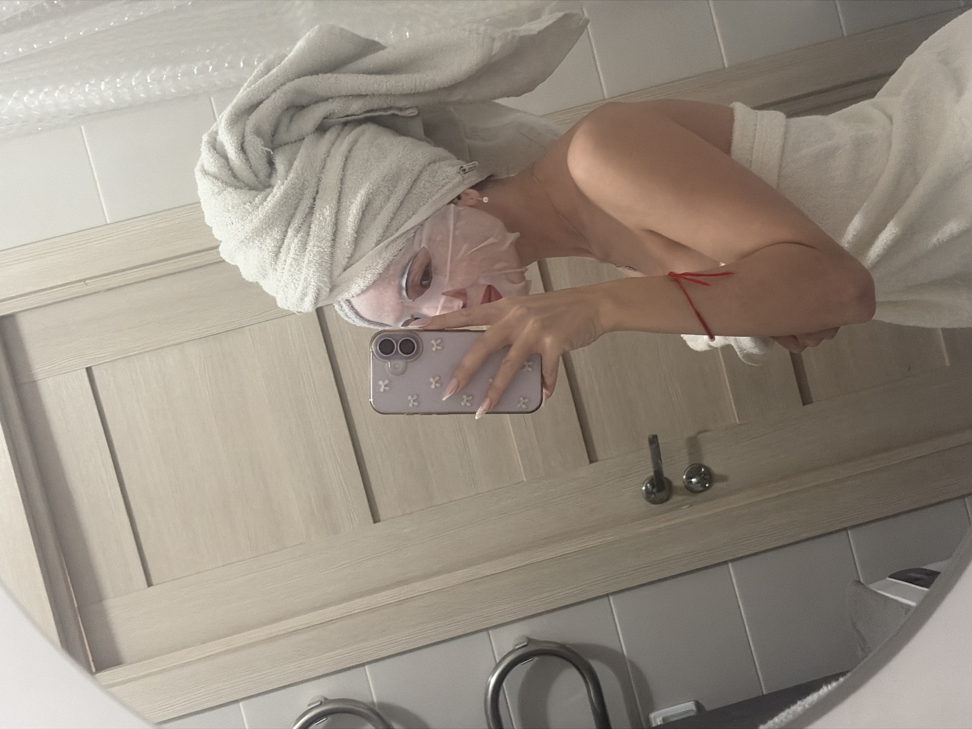 A woman with a white towel wrapped around her head and a white sheet mask on her face is taking a selfie in a bathroom mirror. She is holding a purple iPhone with her right hand. She is wearing a white towel wrapped around her torso. A red string bracelet is on her left wrist. The bathroom has white tiles and a light wood-colored vanity with silver-colored handles. The vanity has a shelf with two faucet handles visible. The location appears to be a bathroom in a residence, and there is no indication that it is in Saint Petersburg, Russia.