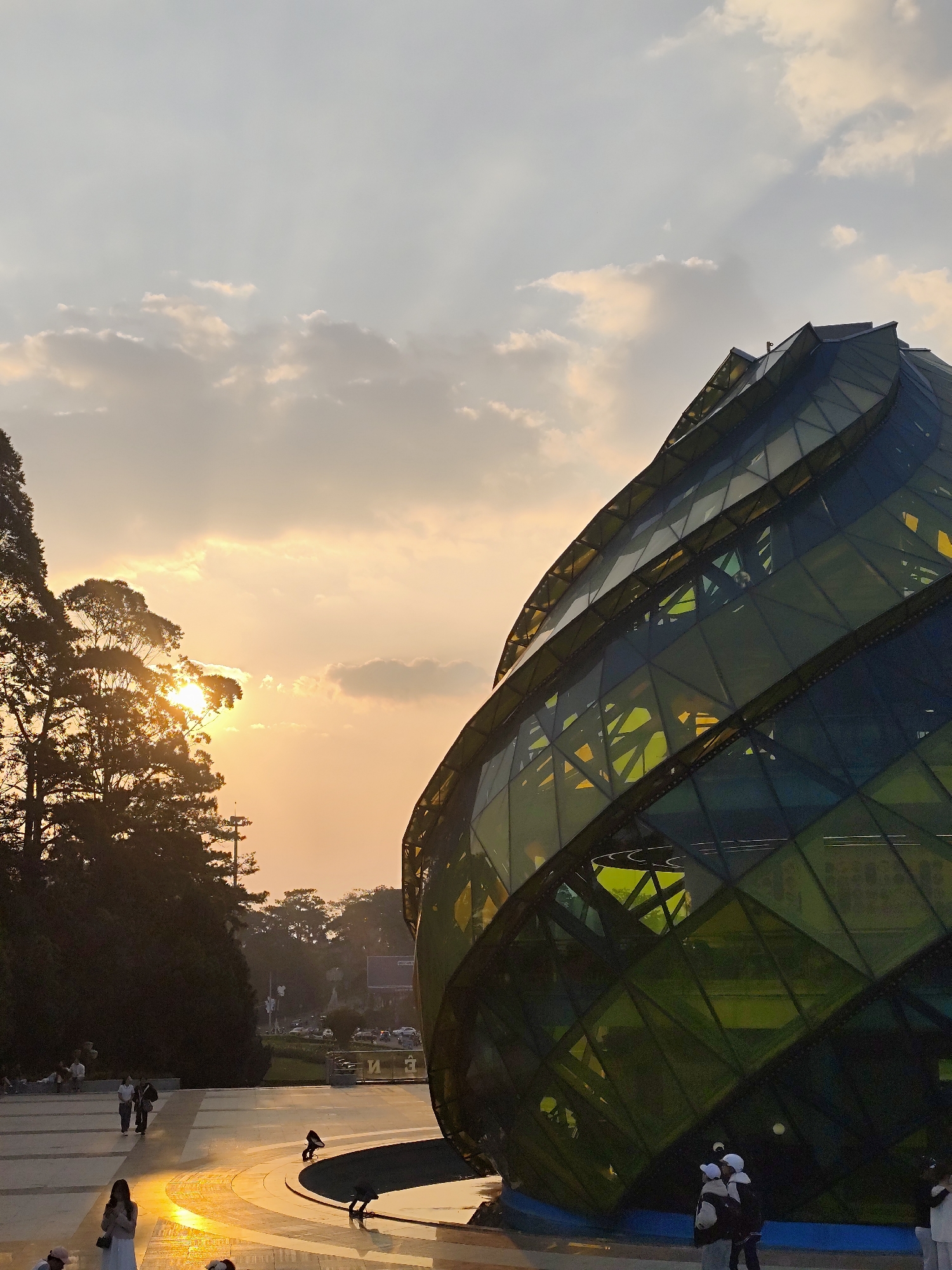 The image depicts an outdoor scene at sunset at the Lam Dong Convention Center (also known as the Da Lat Glass House) in Da Lat, Vietnam. The prominent subject is a large, ovoid-shaped building occupying the right half of the frame, constructed from numerous green and dark blue triangular and quadrilateral glass panels forming a spiraling, faceted exterior. The setting sun, a bright orange-yellow orb, is visible on the left behind the silhouette of a tall tree, casting warm light across the sky with scattered clouds and illuminating a paved plaza in the foreground.
On the plaza, several individuals are present. Near the bottom center, a person in a white dress stands, illuminated by the sun's low angle. Further back, two figures walk away from the viewer. At the base of the glass structure, a group of people, some wearing white head coverings, stands near a low, circular water feature with integrated spotlights. Additional figures are visible seated or standing in the shaded background area to the left, beneath more trees. The environment is a public park or plaza with cultivated trees and paved walkways.