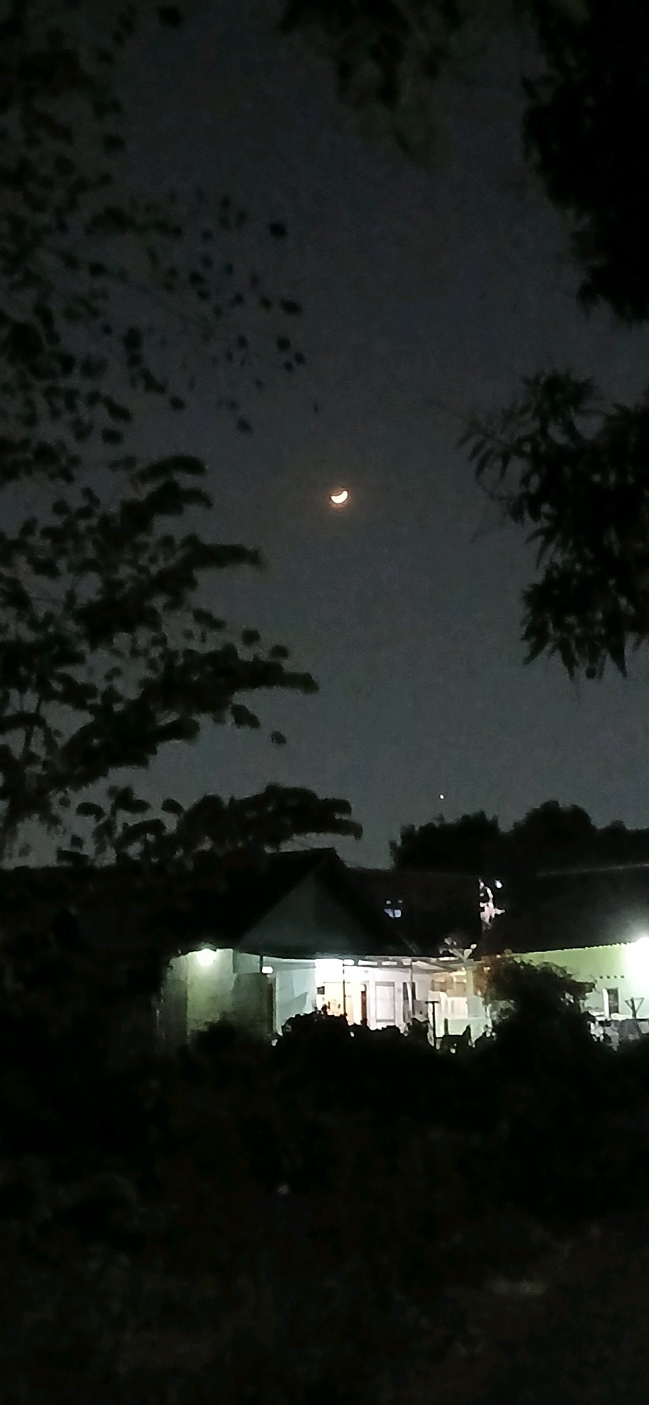The image depicts a night scene with a crescent moon visible in the dark sky. In the foreground, silhouetted trees frame the view of a house. The house appears to be illuminated from within, with lights showing through windows. The surrounding area is dark and appears to be covered in vegetation or foliage, suggesting a semi-rural or residential setting. The location is identified as Sutawinangun, Indonesia. There are no people visible in the image.