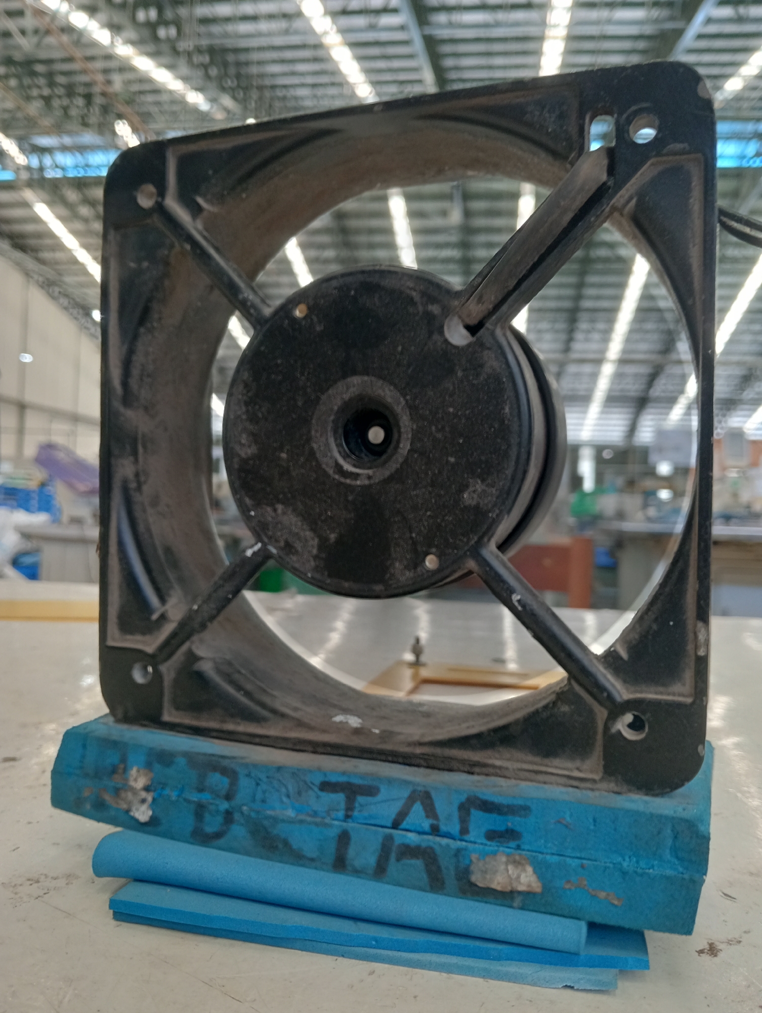 A dusty, black, square fan is propped up on a blue, rectangular block that has black markings and some white scuff marks. Beneath the blue block are two flat, light blue foam pads. The fan has a black circular motor in the center and four black supports connecting the motor to the outer square frame. The background shows a large indoor space with a high, corrugated metal ceiling with rows of lights. There are various objects and structures visible in the background, including some purple and blue items on shelves or tables in the mid-ground and some blurry industrial equipment further back. The scene appears to be an interior of a warehouse or factory.