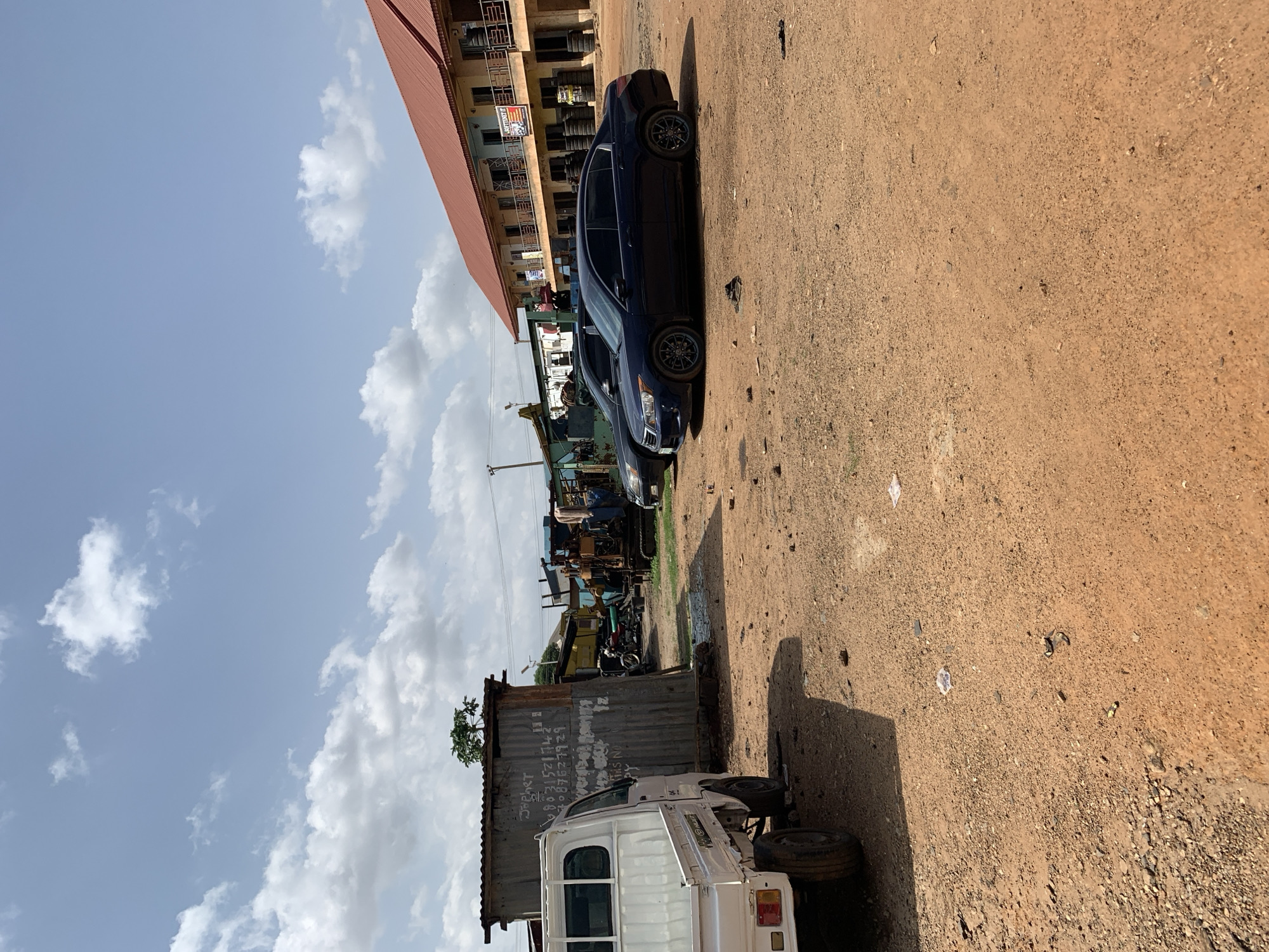 The image depicts an outdoor scene in Rafin Par, Nigeria, under a bright, partly cloudy sky, suggesting it is daytime. The foreground is dominated by a dirt or gravel ground.
The primary subjects are vehicles and buildings. A dark blue sedan is parked prominently in the mid-ground, facing to the left. Behind it, a white truck is partially visible, facing to the right and appearing to be a commercial vehicle. Further in the background, there are various structures, including a building with a red roof and light-colored walls, likely a shop or a residence, with rows of tires stacked outside. There are also what appear to be shipping containers and other assorted industrial or commercial equipment.
The scene suggests a commercial or industrial area, possibly a marketplace or a workshop, given the presence of stacked tires and other equipment. The overall atmosphere is that of an active, perhaps busy, location.
Some text is visible on one of the shipping containers, although it is partially obscured and difficult to read clearly. It appears to include numbers and some letters, possibly identifying markings or contact information. The ground is uneven and appears to be a dirt lot, with some scattered debris. The lighting suggests it is a sunny day, with shadows cast by the vehicles and structures.