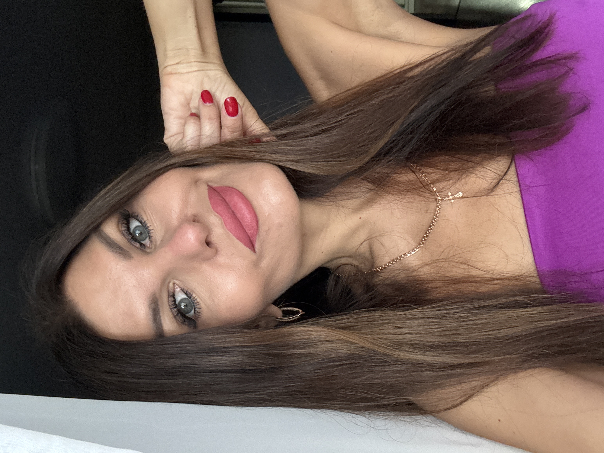 A woman with long, dark brown hair and blue eyes, wearing pink lipstick and a purple top, poses for a selfie. Her left hand is resting on her head, and her right arm is extended behind her. A gold necklace with a cross pendant is visible around her neck. The background is dark, with a white curtain on the left side of the image. The scene appears to be indoors, in a domestic setting. There is no information in the image to suggest a location in Chaikovsky, Russia.