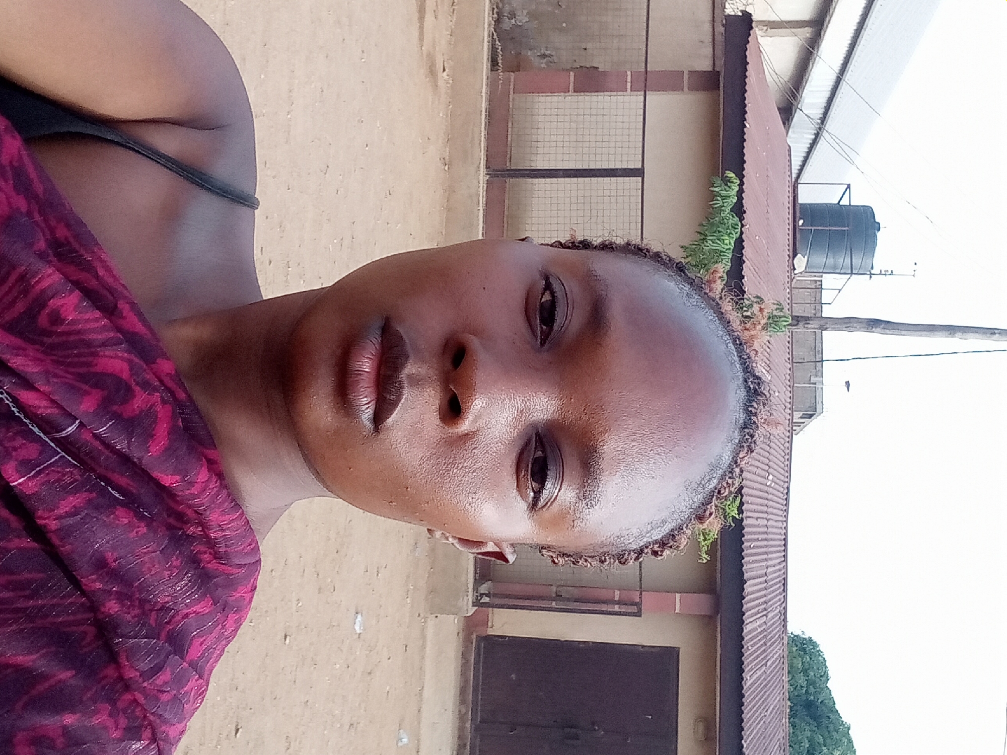 A medium shot of a dark-skinned woman with a shiny face, wearing a dark purple and pink patterned top and black tank top straps, against a backdrop of a beige-colored building with a brown corrugated roof. There's a black water tank on the roof, and some greenery is visible. The setting appears to be Sabon Tasha, Nigeria.