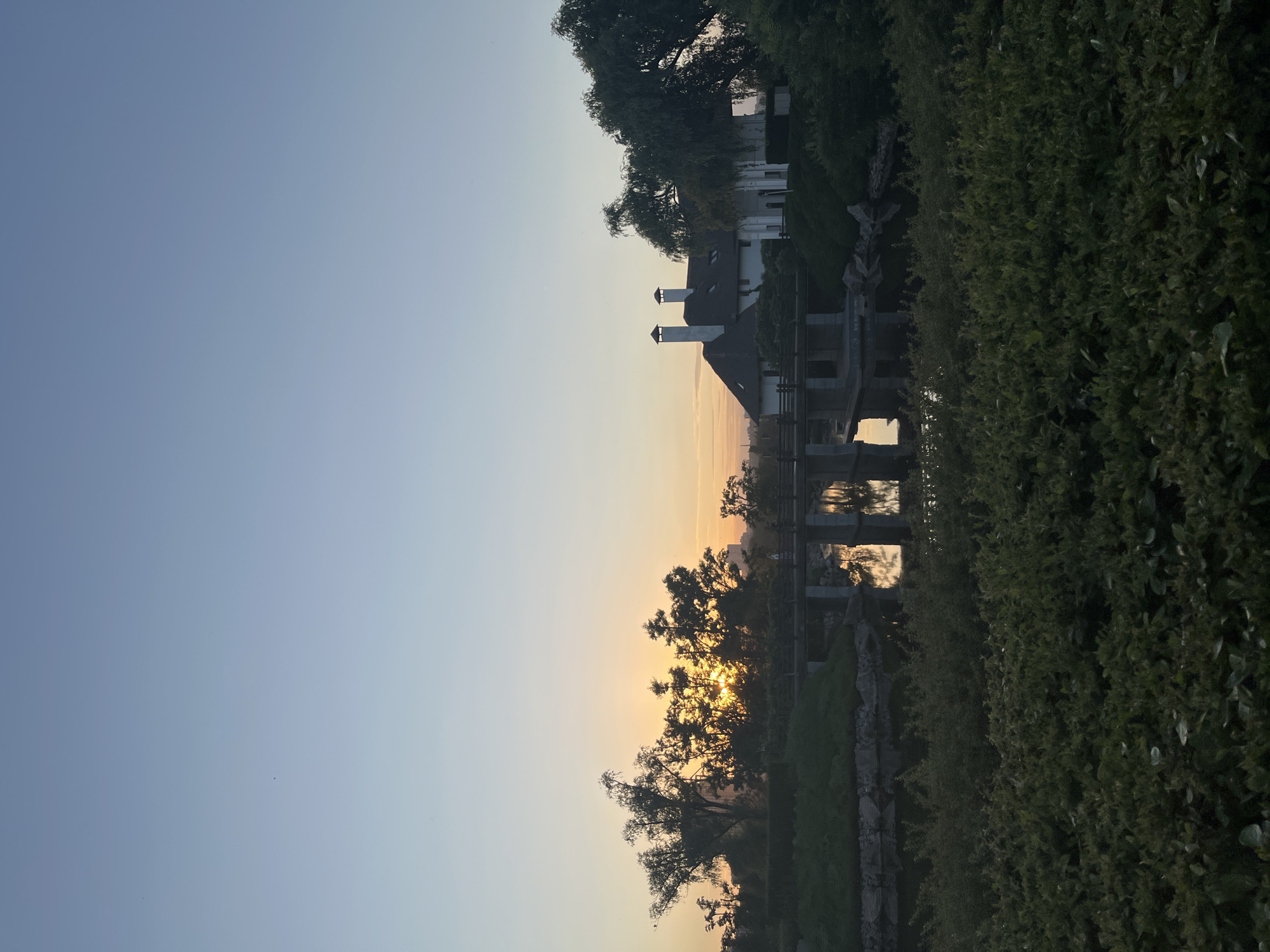 The image depicts a tranquil park scene at dawn or dusk, likely in Dnipro, Ukraine, given the context provided. The sky is a gradient of pale blue and orange, indicating the low angle of the sun which is partially obscured by a dense cluster of trees on the left side of the frame. The sun's rays create a glowing effect through the branches. In the mid-ground, a modern-style building with a white facade, dark roof, and two prominent chimneys stands to the right. A bridge with stone pillars and a wooden railing spans a calm body of water in front of the building. The water reflects the bridge, the building, and the surrounding greenery. The foreground is dominated by lush, dark green bushes and foliage. The overall atmosphere is serene and natural.