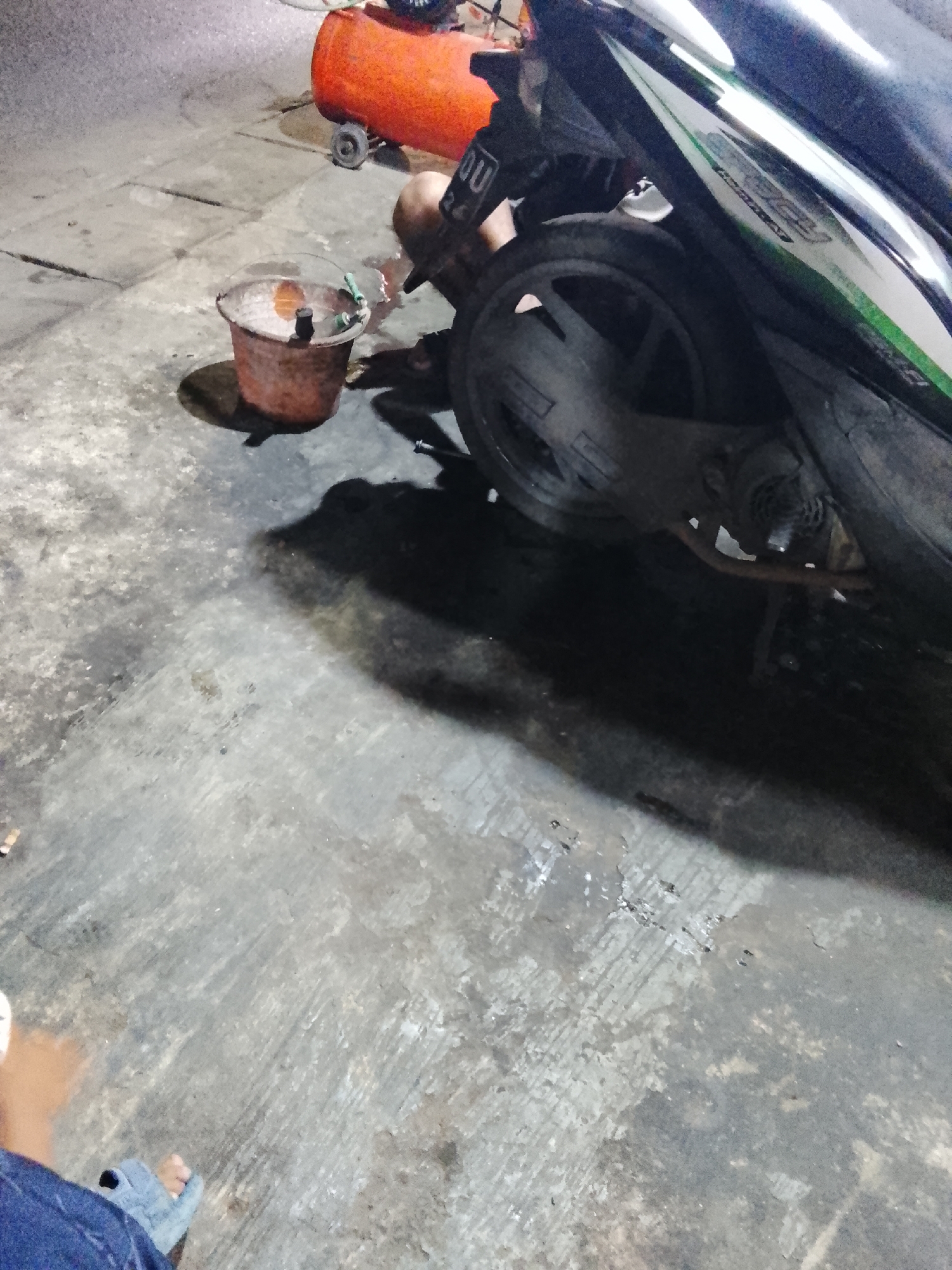 The image depicts an outdoor scene, likely in an urban or semi-urban environment, possibly in Jayasampurna, Indonesia. In the foreground, a portion of a person's legs and feet, wearing blue jeans and sandals, is visible on a concrete ground surface. The ground is rough and appears weathered, with patches of oil or water stains.

To the left of the frame, a small, rusted metal bucket is present on the ground, containing a liquid, possibly oil or water, and a floating object. Behind the bucket, an orange air compressor with wheels is visible. In the mid-ground and right side of the image, a motorcycle is parked, with its rear wheel and part of its body visible. The motorcycle has a black fairing with a green and white decal. A person's legs and torso, wearing dark clothing, are partially visible near the motorcycle, possibly engaged in some activity related to it or the air compressor. The overall lighting suggests it might be evening or nighttime, with artificial light sources casting shadows. The environment appears to be a workspace or a service area.