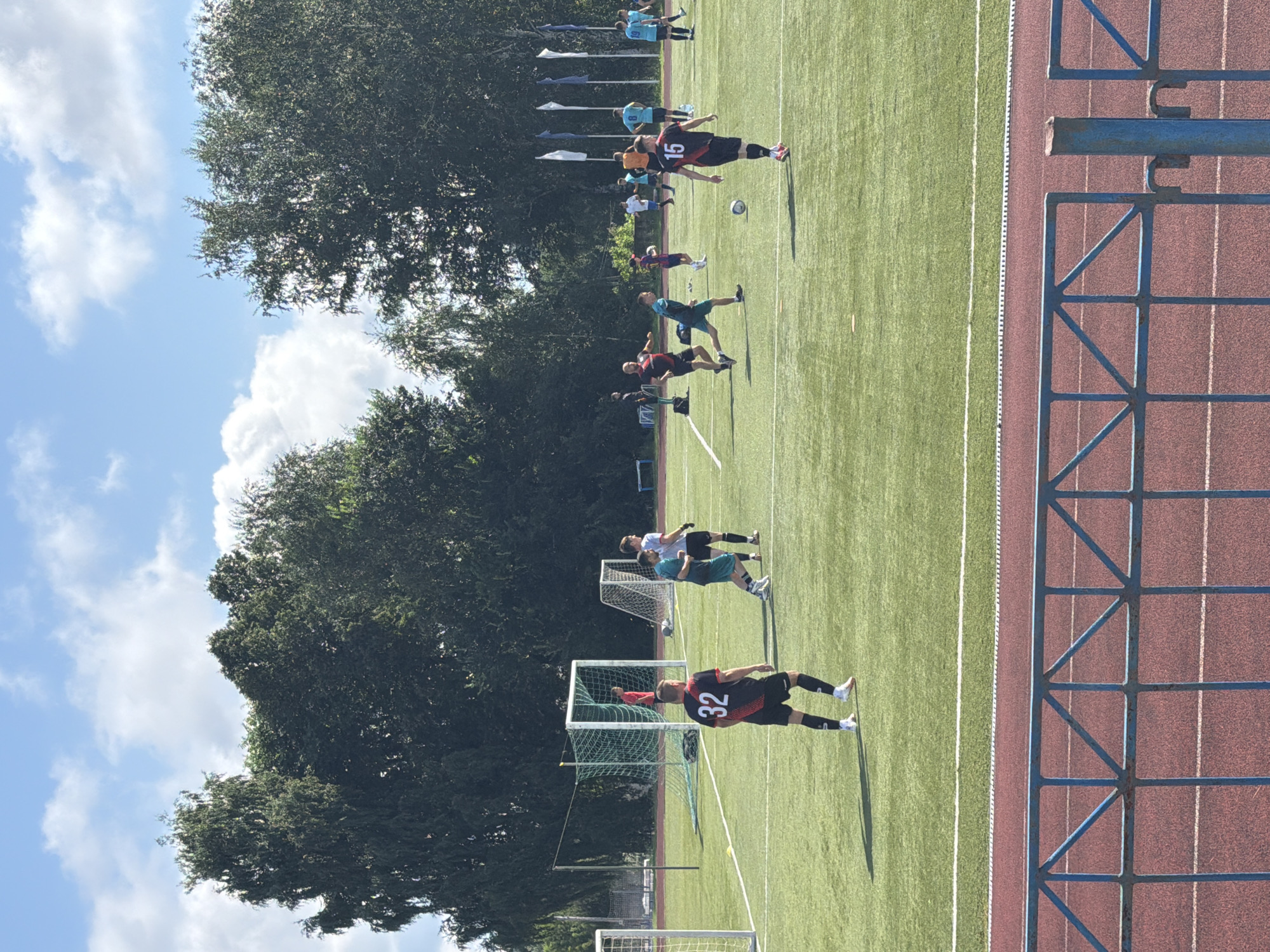 The image shows a soccer game taking place on an artificial turf field in Chaikovsky, Russia. In the foreground, a player wearing a black and red jersey with the number 32 on the back is standing on the field, looking towards the right. Behind him, several other players are engaged in the game. The field is marked with white lines, and there are soccer goals visible at either end. The surrounding area is bordered by a red track and a blue fence. Trees and a cloudy sky form the background.