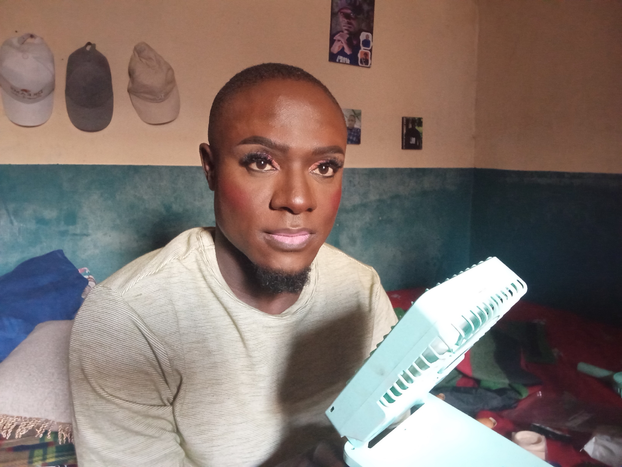 The image shows a medium shot of a person wearing makeup, including foundation, blush, eyeshadow, mascara, and lipstick. The person has short, dark hair and is looking directly at the camera. They are wearing a light-colored, ribbed t-shirt. To the left of the person, on the wall, are three baseball caps hanging: one white, one gray, and one beige. Above them, on the wall, are two small framed pictures. To the right of the person, in the foreground, is a light blue portable fan. The background consists of a wall painted in two tones, with a teal band across the middle and a lighter color above. The environment appears to be an indoor setting, possibly a room in Nigeria, given the reference to Rukuba Road. The overall impression is a personal portrait with elements of the surrounding environment visible.