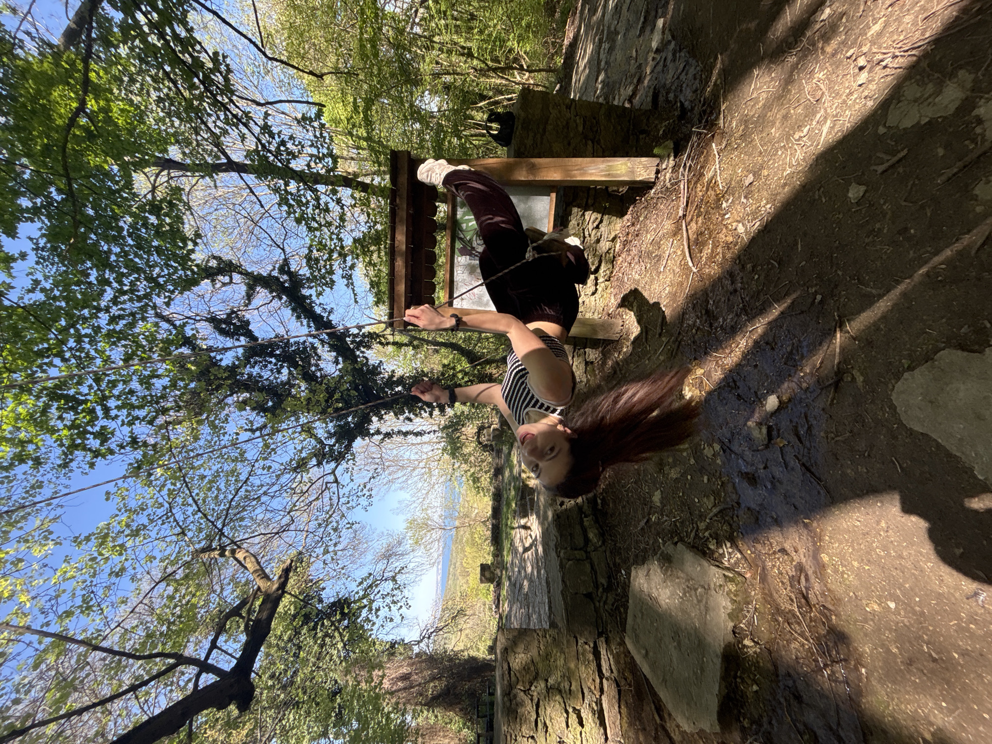 A female is hanging upside down from a rope swing in a wooded area. She is wearing a striped crop top, dark pants, and white sneakers. Her long brown hair is hanging down towards the ground. She is holding onto the rope swing with both hands. The background shows trees, a blue sky, and a stone structure. The ground is dirt with some stones and branches. The location is likely a wooded area in or near Varna, Bulgaria, given the landscape and architectural elements. There are trees, paths, and structures typical of parks and recreational areas in that region.