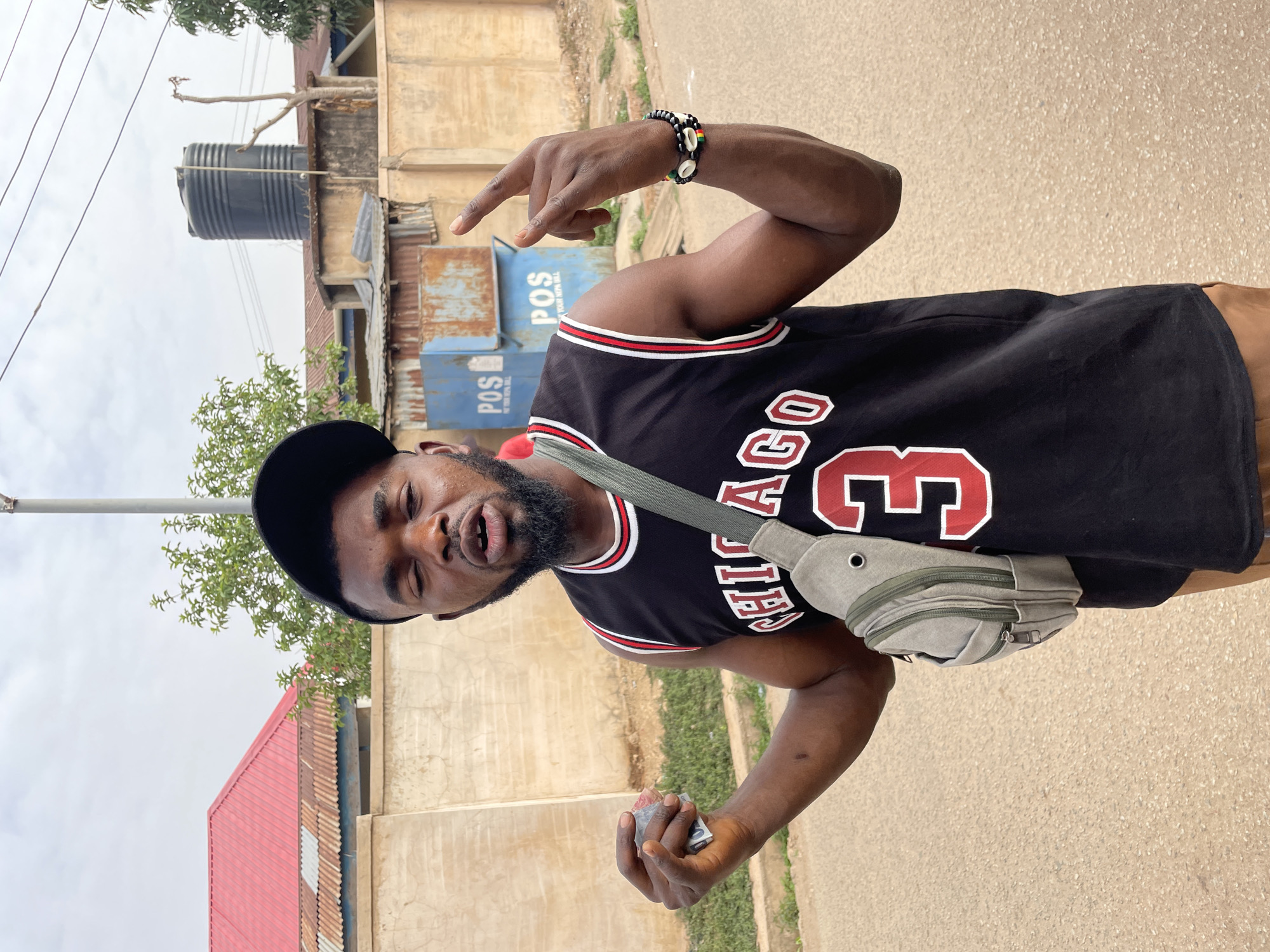 A man is outdoors on a paved surface in Jos, Nigeria. He is wearing a black Chicago Bulls jersey with the number 3, a black baseball cap, and a bracelet made of beads and shells on his right wrist. He also has a small gray bag across his chest and is holding money in his left hand. The man is making a hand gesture with his right hand.
Behind him are buildings of corrugated metal sheets. The sky is overcast, and there is green vegetation in the scene. A water tank is visible on the rooftop of one of the buildings. Stacks of POS containers are visible behind the buildings. A street pole rises above the man's head.