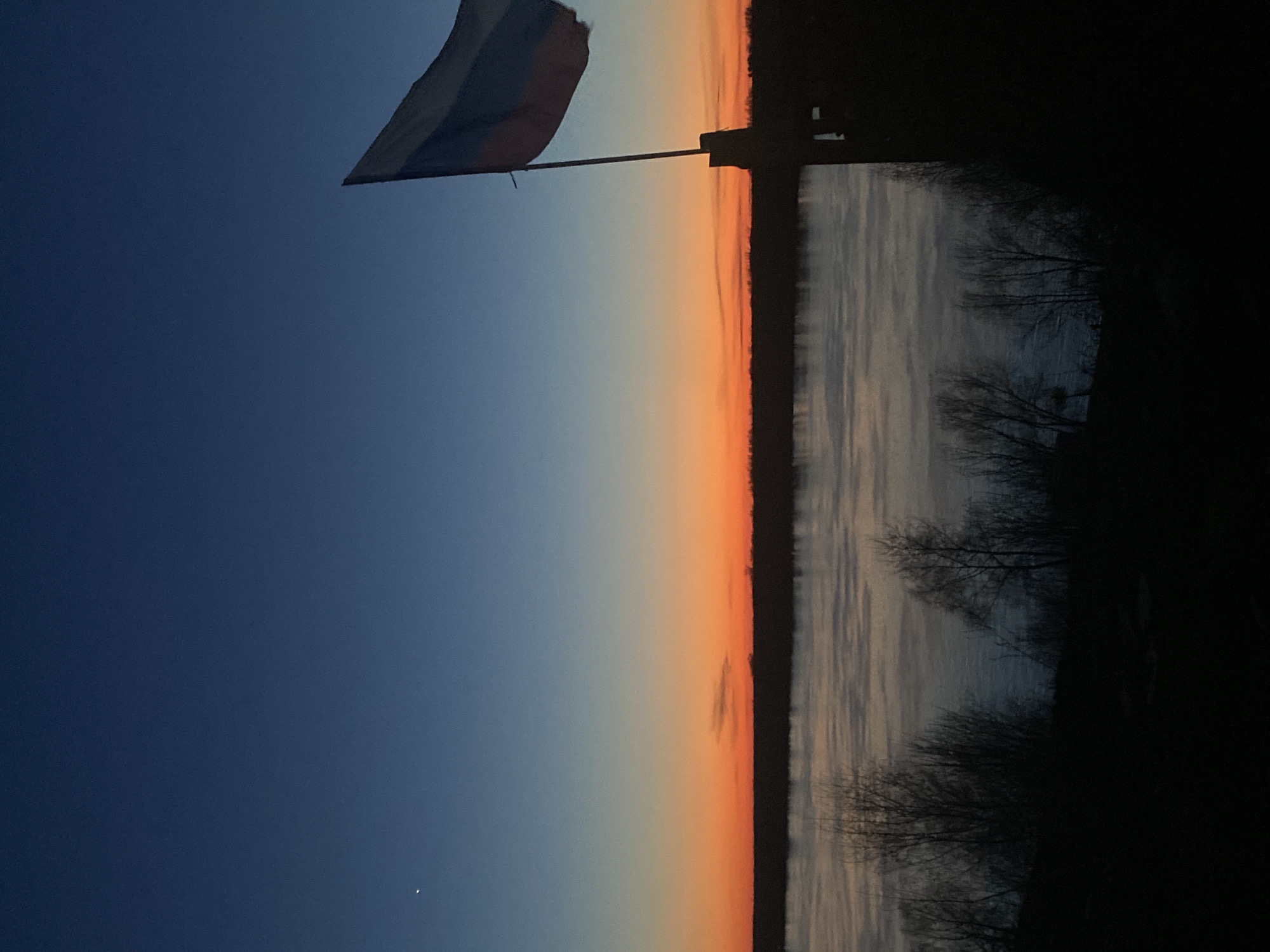 The image depicts a landscape at dusk or dawn, possibly in Ольховка, Russia, given the Russian flag on a pole in the upper left. The sky transitions from dark blue on the left to orange near the horizon, possibly indicating a sunset or sunrise. A body of water, possibly a river or lake, reflects the sky, with trees lining the shore on the right side, which is shrouded in darkness. The flag is the prominent object, suggesting a location of significance, possibly a governmental building or public space. There are no visible people, actions, or interactions in the scene. The general environment is serene and quiet, with a natural landscape setting near water. A small point of light, possibly a star or planet, is visible in the dark blue part of the sky, indicating a clear atmosphere.