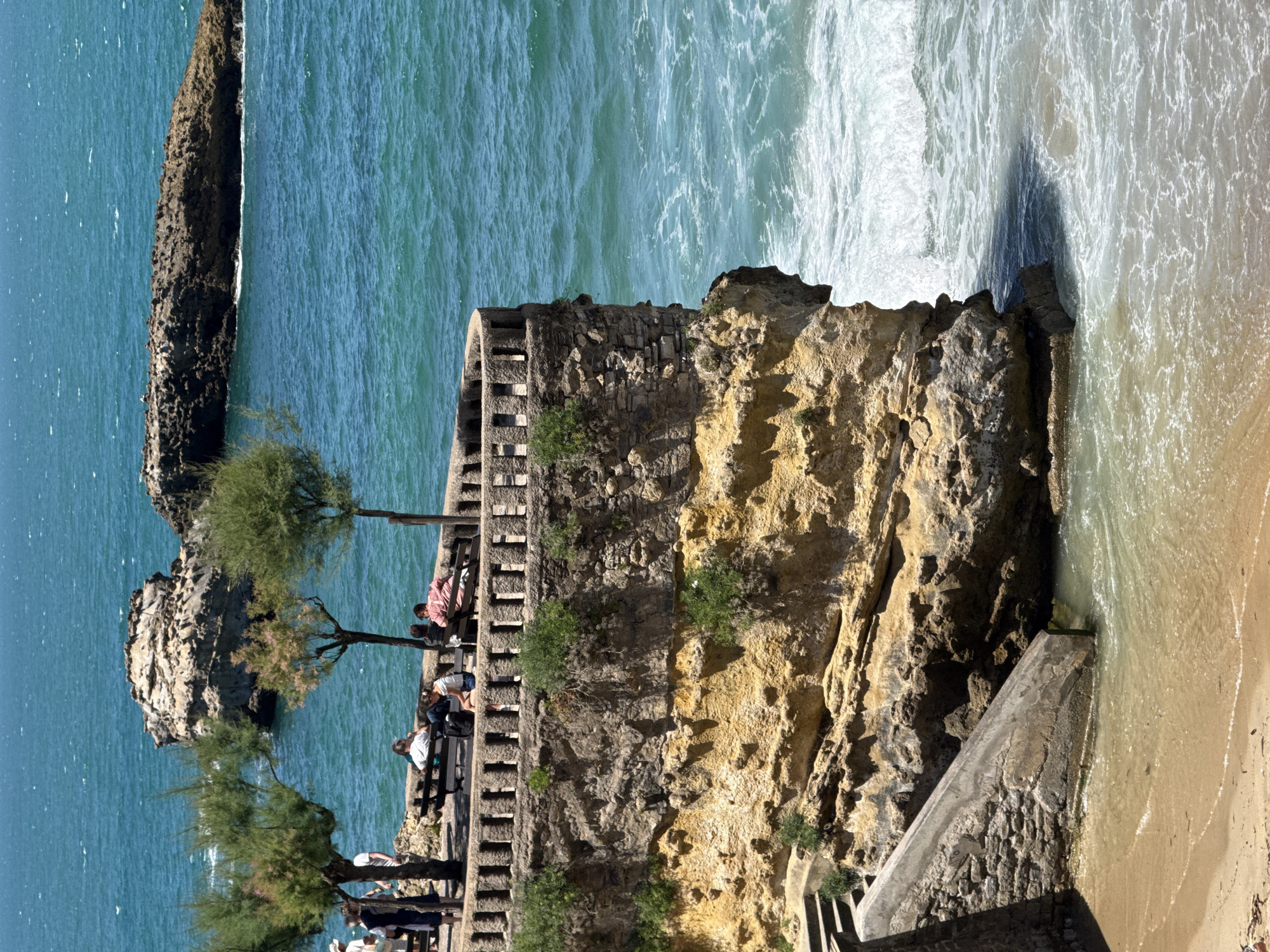 This image captures a sunny day at the Rocher de la Vierge in Biarritz, France. A stone promenade, adorned with a decorative railing and a few sparse trees, overlooks the turquoise ocean. Several people are seated on benches along the promenade, enjoying the view. Some are engaged in conversation, while others are simply relaxing. Below the promenade, a rocky cliff face descends to a small sandy beach where gentle waves lap the shore. In the background, a prominent rock formation rises from the sea, adding to the picturesque coastal scenery. The overall impression is one of a peaceful and scenic day at the seaside.