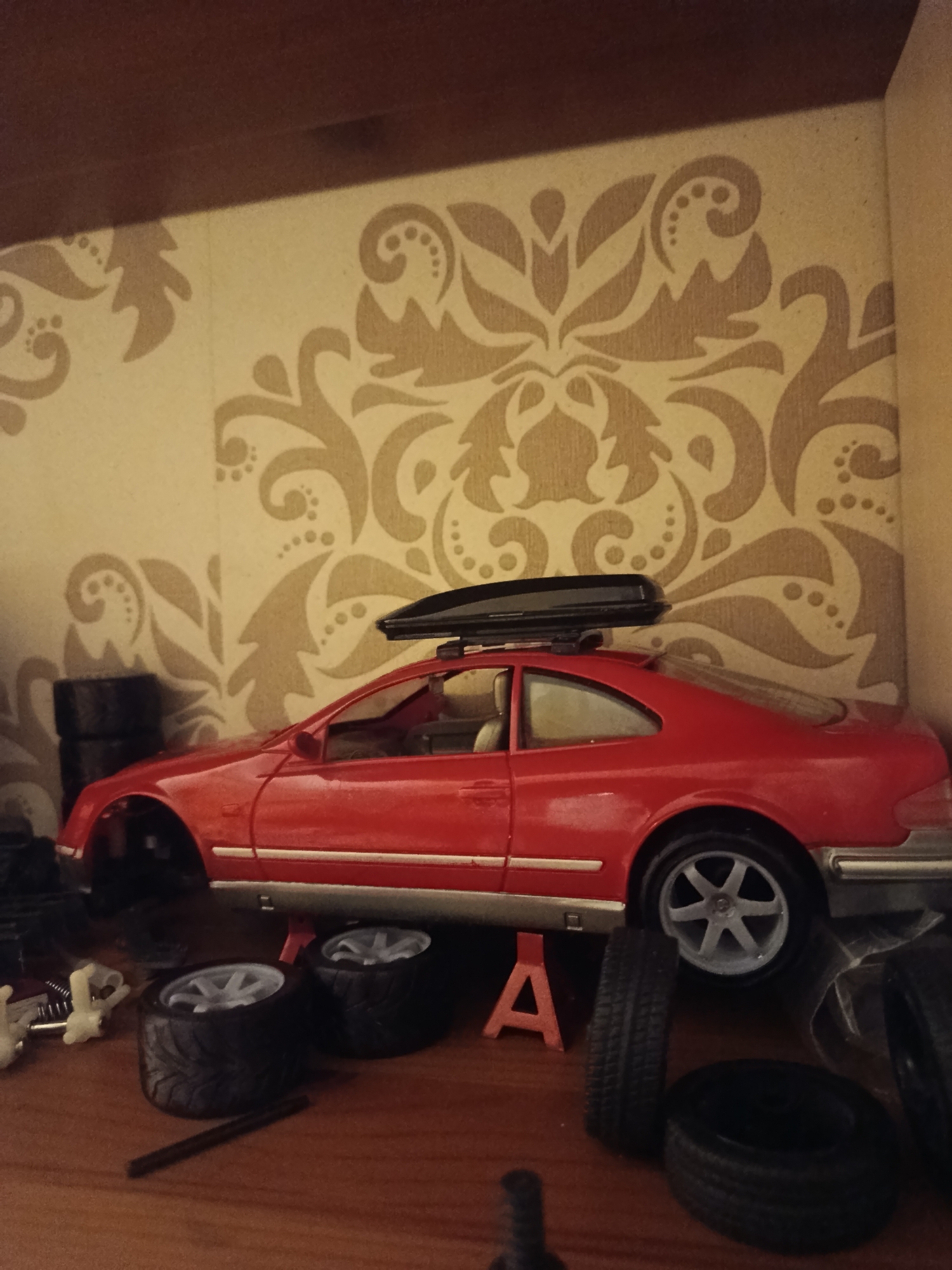 The image shows a close-up view of a red toy car, possibly a Mercedes-Benz, with a black roof rack. The car is elevated on red jack stands, suggesting it is undergoing maintenance or repair. Several spare tires are scattered around the car, some with silver rims. In the background, a wall is visible with a light-colored wallpaper featuring a damask pattern in a beige hue. A wooden shelf or surface is also present at the top of the image. The scene appears to be indoors, possibly in a garage or workshop. There are no people present in the image. The image is taken from a slightly elevated angle. The context of Sertolovo, Russia is not discernible from the visual information provided in the image.