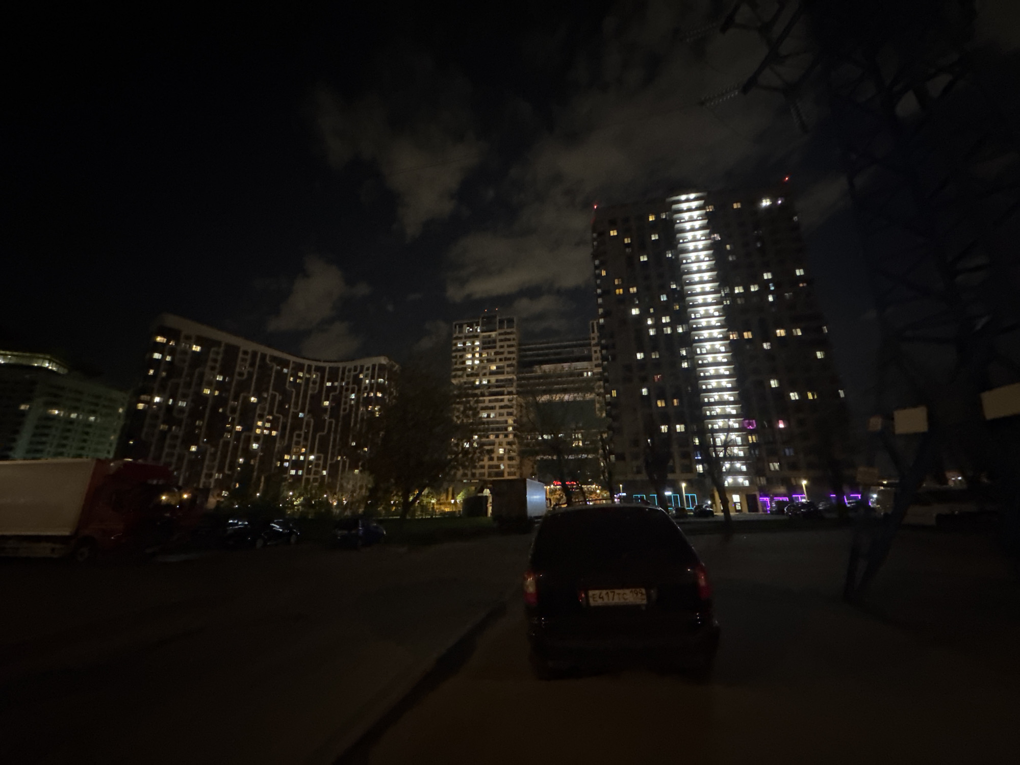 At night in Moscow, Russia, several tall, modern apartment buildings with numerous illuminated windows stand against a dark sky with scattered clouds. A parked black car faces the viewer, its license plate reading "E417TC 199." A large white truck is parked to the left, partially obscured by darkness. Several other parked vehicles are visible in the background. In the lower right corner, a dark figure stands near a large, shadowy metal structure, possibly a utility pole. The scene appears to be a street or parking area.