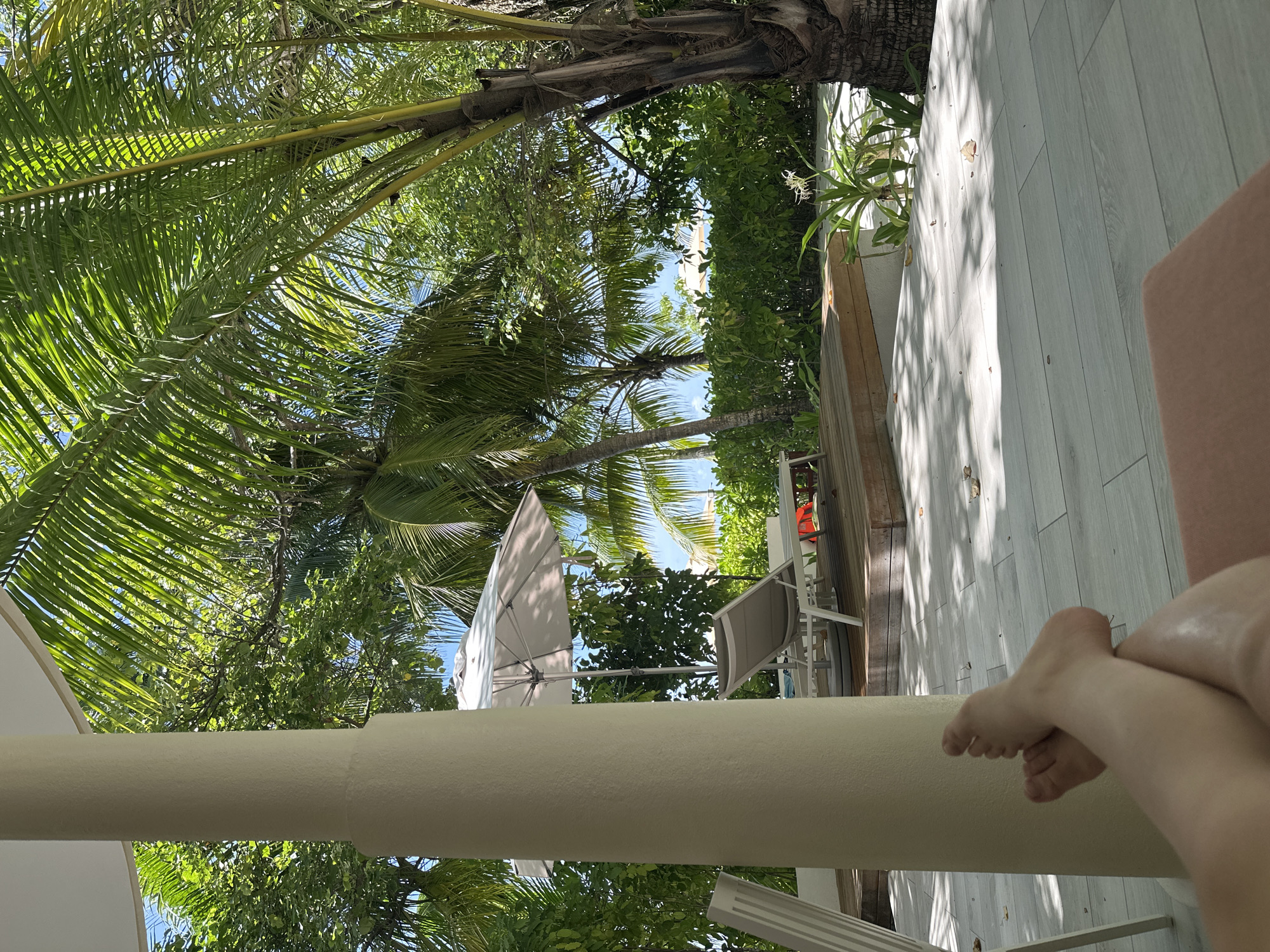 In Halaveli, Maldives, a close-up shot captures a person's bare feet resting in the foreground, crossed at the ankles, as they recline on a cushioned lounge chair. The viewpoint is from a covered patio with a cream-colored pillar to the left. Beyond the patio, a wooden deck features two white lounge chairs and a large white umbrella. Lush green foliage and palm trees dominate the background, with a glimpse of the bright blue sky and the ocean peeking through. Sunlight filters through the palm fronds, casting dappled shadows on the pale tiled floor. The scene conveys a relaxed and tropical atmosphere.