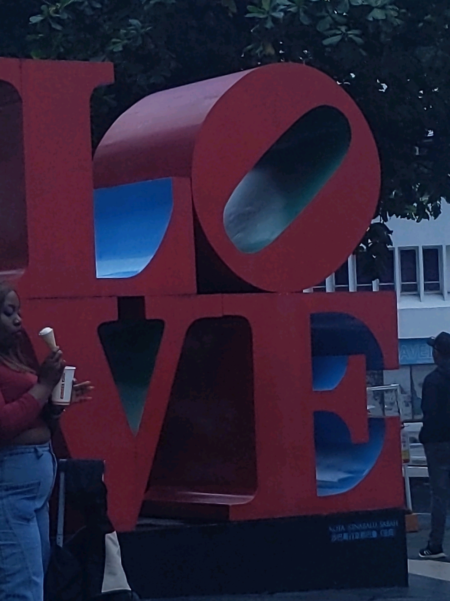 A large, red, three-dimensional sculpture spelling out "LOVE" is the central focus of the image. The letters are solid and have cut-out sections that reveal a blue background, possibly representing the sky or water.
On the left side of the image, a woman wearing a red top and blue jeans is holding a white cup and a white cone-shaped object. She is looking down at the cup as she holds it.
In the background, to the right of the sculpture, a person wearing dark clothing and a baseball cap is visible. Further in the background, there is a white building with several windows. A sign with the word "AVE" is partially visible.
At the base of the "LOVE" sculpture, there is a black platform with text written on it in a lighter color, which appears to be in both Latin and Chinese characters. The text includes "KOTA KINABALU SABAH" and what looks like a Chinese inscription, "沙巴亚庇市". This indicates the location is Kota Kinabalu, Sabah, Malaysia.
The overall lighting suggests it might be late afternoon or early evening, with a muted, slightly overcast quality to the light. The environment appears to be an outdoor public space, possibly a plaza or park, with trees visible at the top of the image.