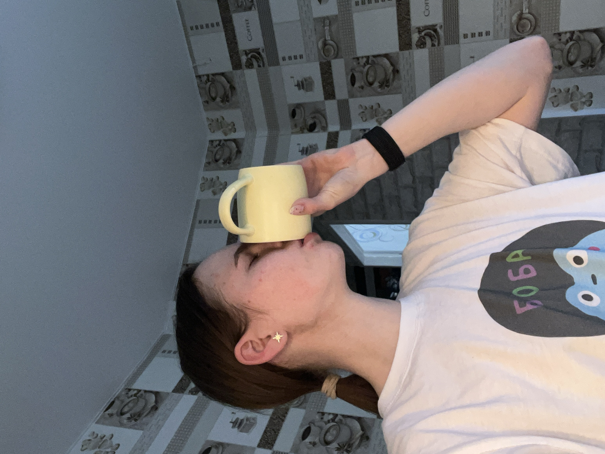 The image shows a woman indoors, holding a yellow mug to her forehead. She is wearing a white t-shirt with a cartoon print and has dark hair pulled back in a ponytail. She has a star-shaped earring in her left ear. The background features a wall with patterned tiles showing coffee-related images. A white table is partially visible. The precise location within the city of Chaikovsky, Russia cannot be determined from the image.