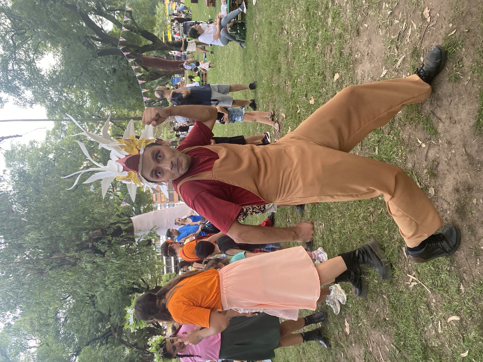 A performer in costume, wearing a red shirt and tan overalls with a large feathered headdress, poses with raised fists at a public event. The performer is on grass and looks towards the camera. There are many people behind the performer. Trees are visible in the background. This location is in Buenos Aires, Argentina.