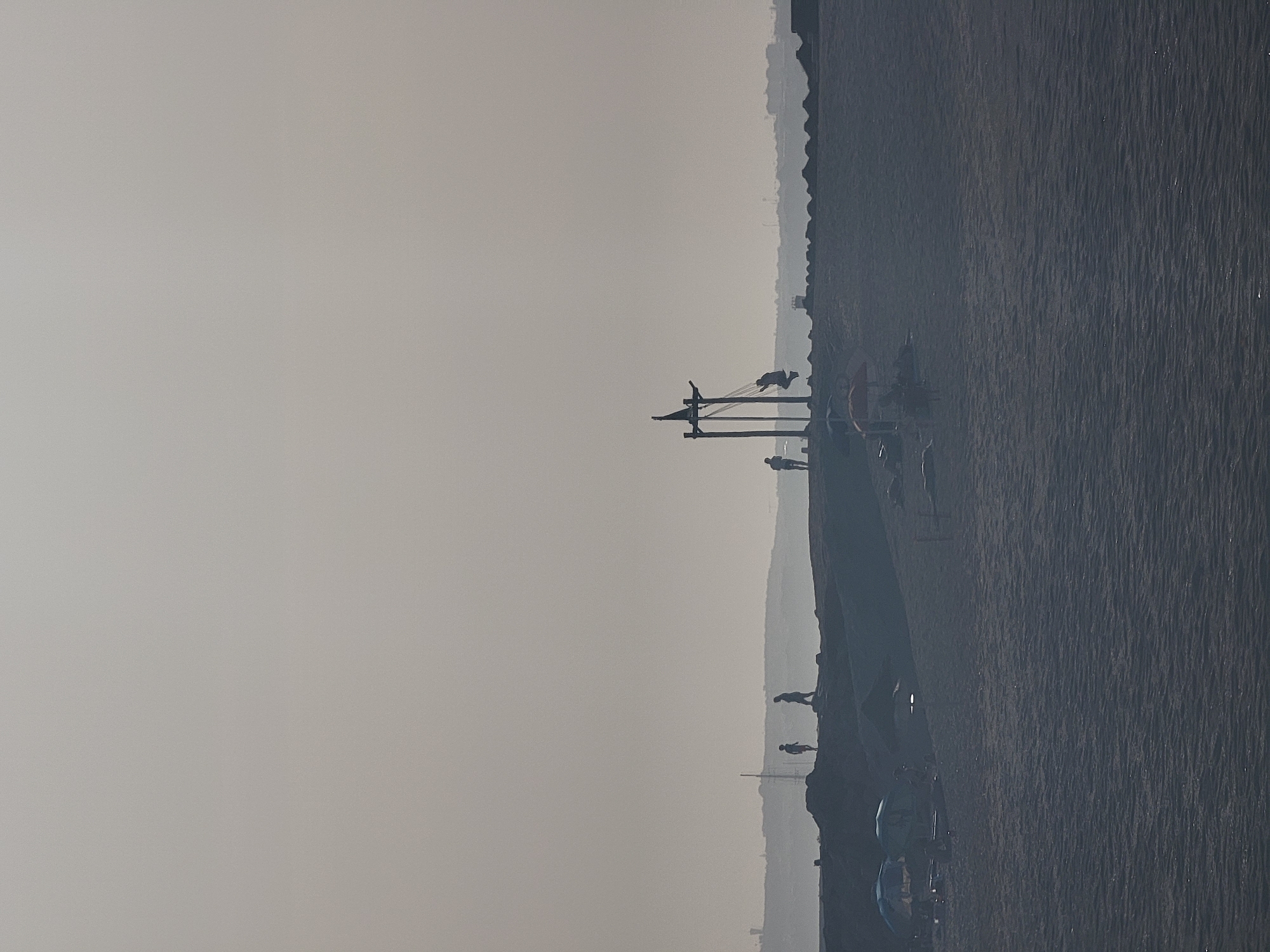 The image depicts a hazy, overcast day at a beach in Quarteira, Portugal. In the foreground, dark sand with gentle ripples covers the beach, extending towards the sea. In the middle ground, a dark, elevated promenade or breakwater runs horizontally across the frame. Silhouettes of several people are visible on this elevated area. To the left, two beach umbrellas, one blue and one green, stand on the lower beach. Further back, on the elevated promenade, a man is on a swing attached to a wooden structure resembling a ship's mast, with another person standing nearby. Several other figures are scattered along the promenade. In the far background, a low, undulating horizon is visible with indistinct structures and possibly a distant antenna, all softened by the atmospheric haze. The overall scene conveys a sense of a quiet, possibly off-season, day at the beach.