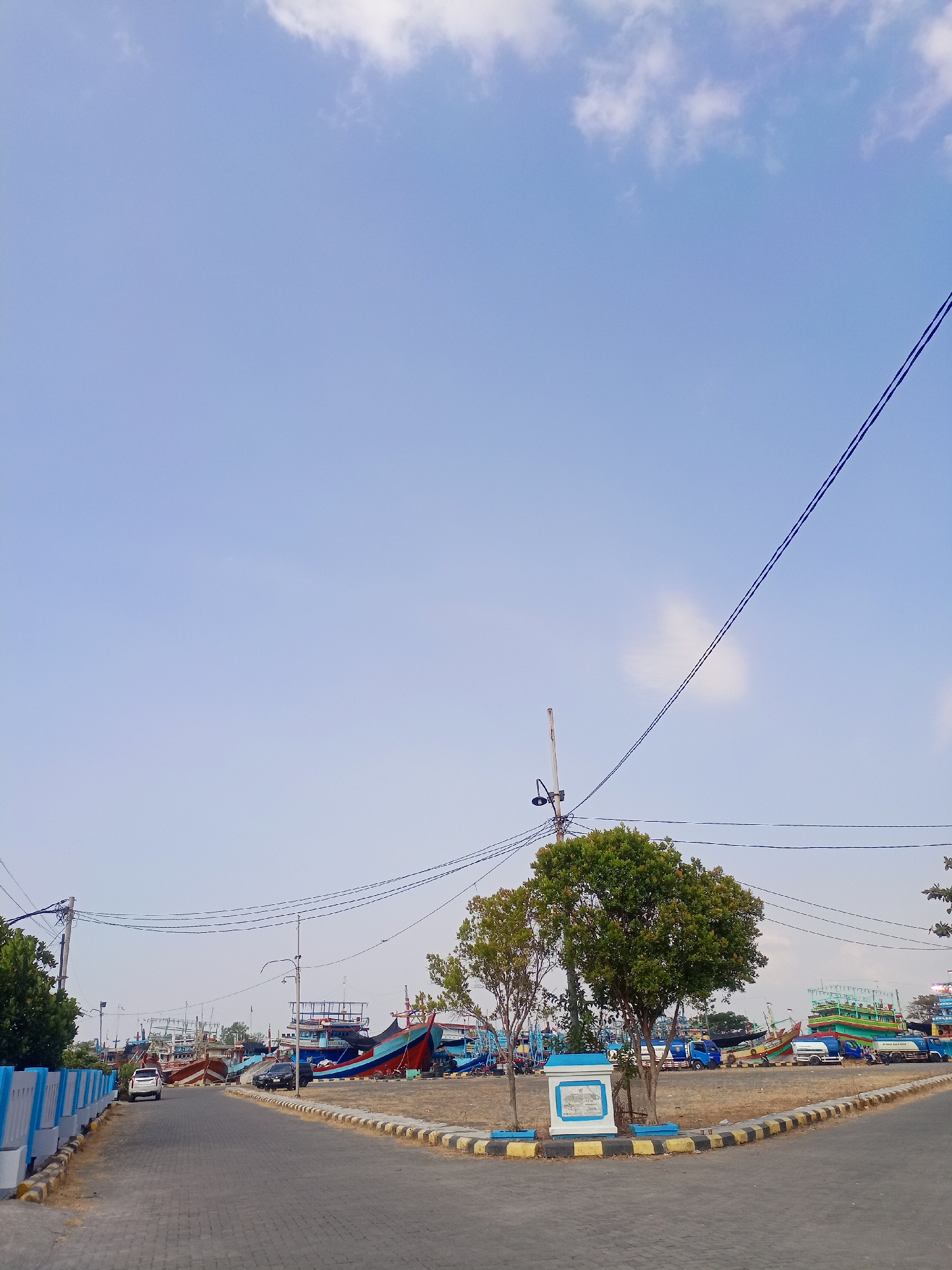 The image depicts a street scene in the city of Juwana, Indonesia, likely at or near the harbor. The foreground shows a paved road with interlocking brick pavers, curving to the right. A low wall with blue and white painted sections borders the left side of the road. In the middle ground, there is a small, decorative monument surrounded by yellow and black striped curbing and two trees. Beyond this, a large number of fishing boats of various sizes and colors are docked or moored. Some of these boats are brightly colored in red, blue, and green. Several vehicles, including cars and trucks, are visible on the road and among the boats. The sky is clear blue with a few scattered white clouds. Power lines are strung across the upper portion of the image. The overall impression is of a busy, working harbor area on a bright, sunny day.