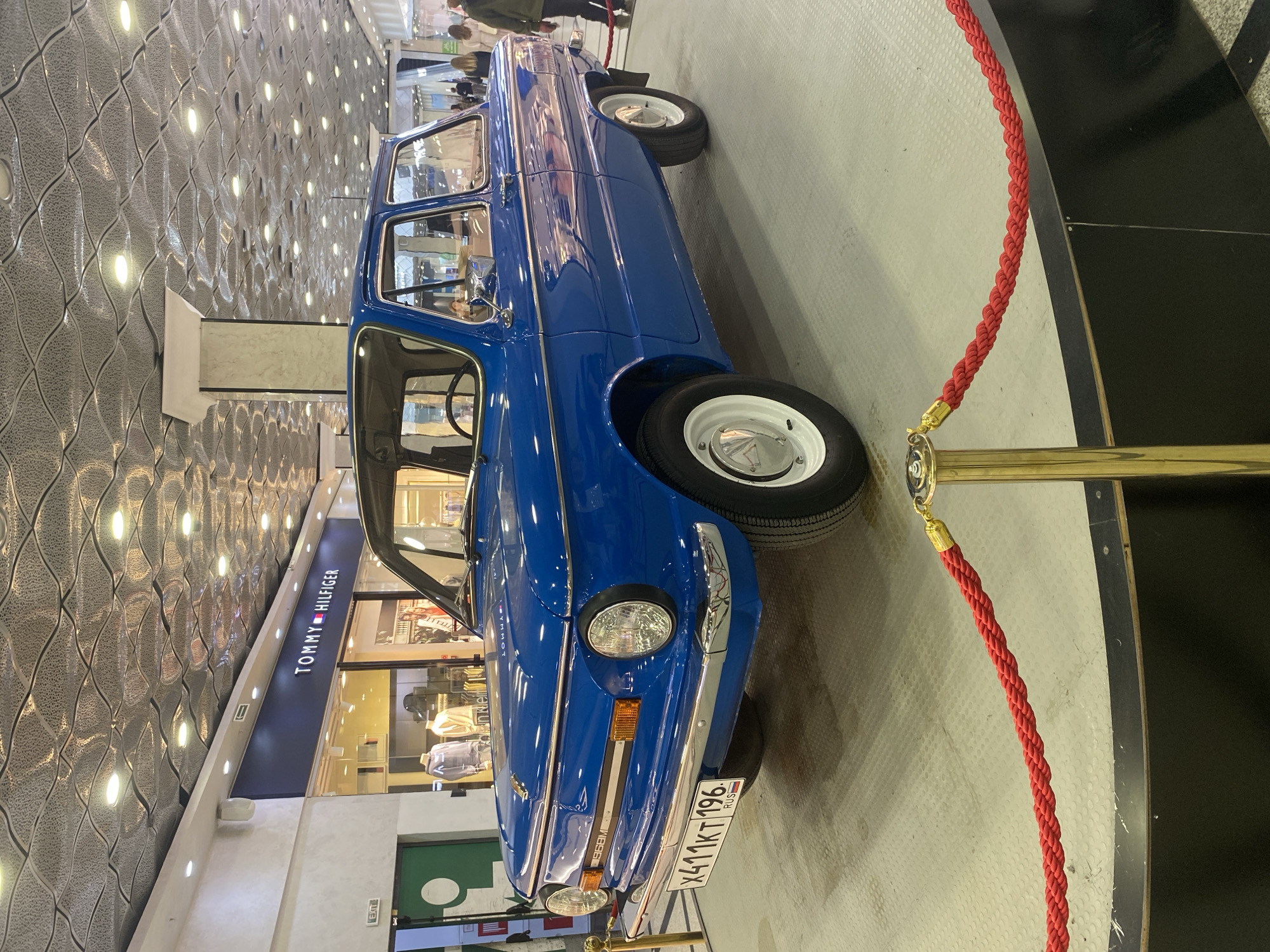 The image depicts a blue vintage car parked near a storefront, likely inside a shopping mall. The car, identified by its license plate as being from Russia (RUS), is parked next to a red rope barrier supported by a gold stanchion. A Tommy Hilfiger store is visible in the background with displays of clothing. Overhead is a patterned ceiling with recessed lighting. This scene takes place in Yekaterinburg, Russia. People can be seen walking in the background.