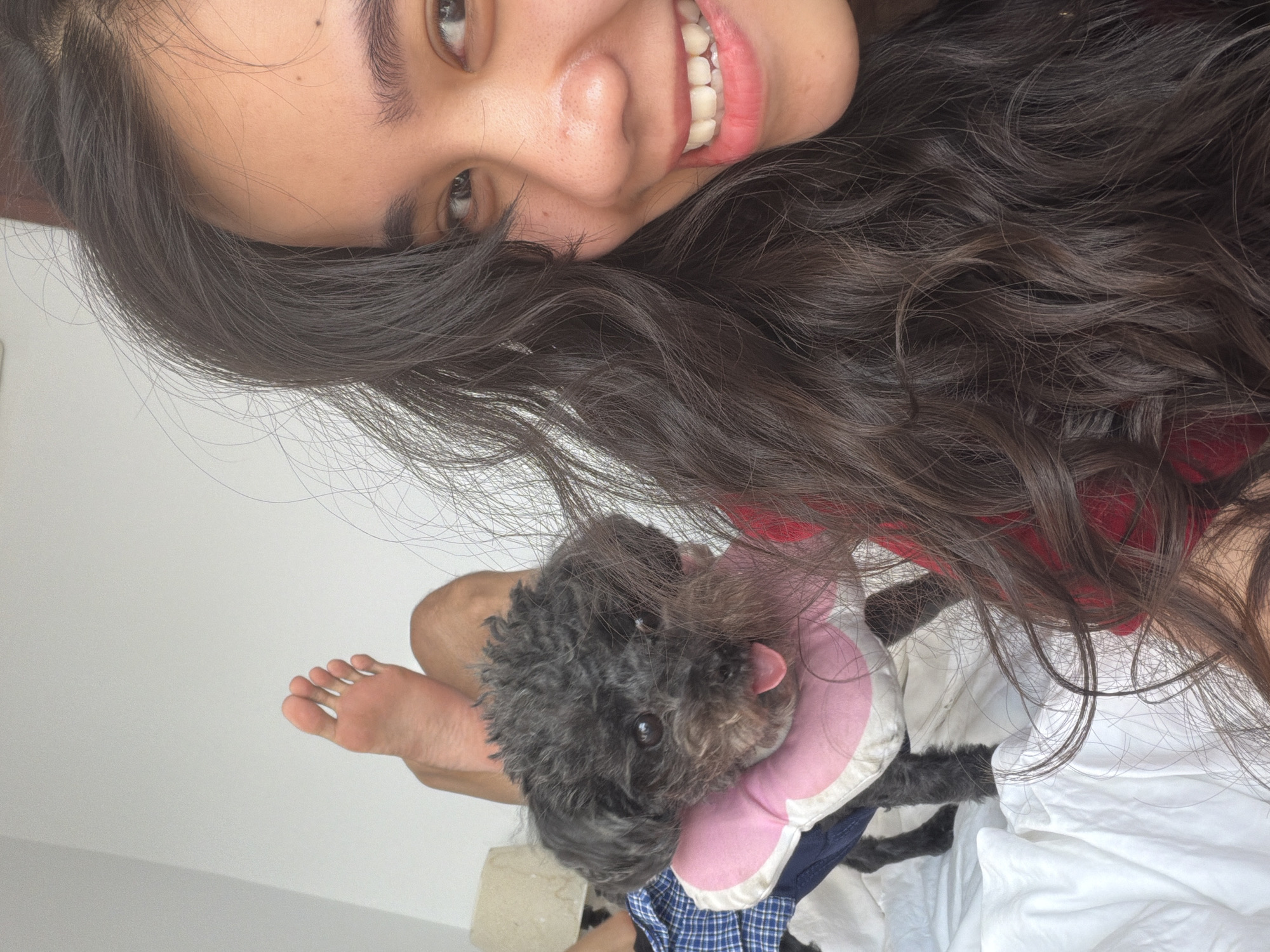 A young woman is photographed at a close, upward angle, smiling and looking towards the camera. She has dark hair, fair skin, and is wearing what appears to be a red top.
Beneath her, a small, dark-haired dog is visible. The dog is wearing a pink and white collar. A foot is visible to the left of the dog. The background includes white bed linens and sections of a white wall.
The location is not explicitly stated as Quy Nhon Ward, Vietnam and cannot be determined from the image alone.