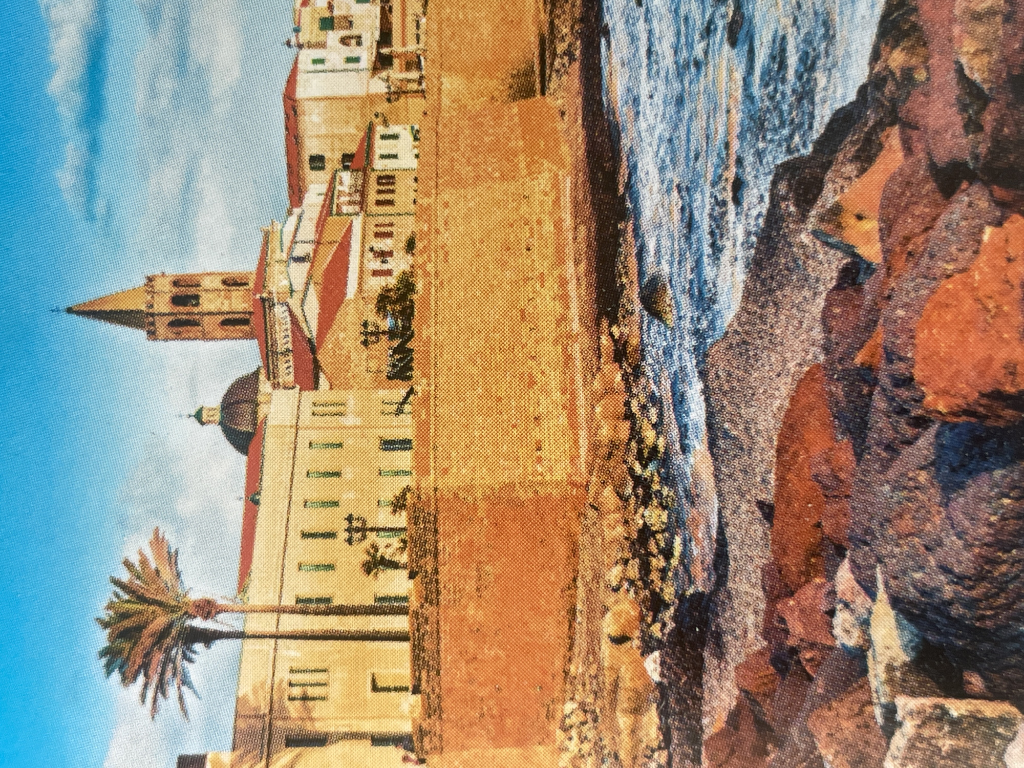 The image depicts a coastal scene with buildings and a body of water. In the foreground, there are rocks and the sea. In the middle ground, there is a stone wall separating the sea from a row of buildings. The buildings are light-colored with red roofs and appear to be of an older architectural style, featuring windows and balconies. A tall bell tower with a spire is prominent among the buildings, as is a dome. A palm tree stands to the left of the buildings. The sky is blue with some clouds. The image is a photograph with a visible dot pattern, suggesting it might be a printed image or a scan. The location is described as the city of Sofia, Bulgaria, which is landlocked and not a coastal city.