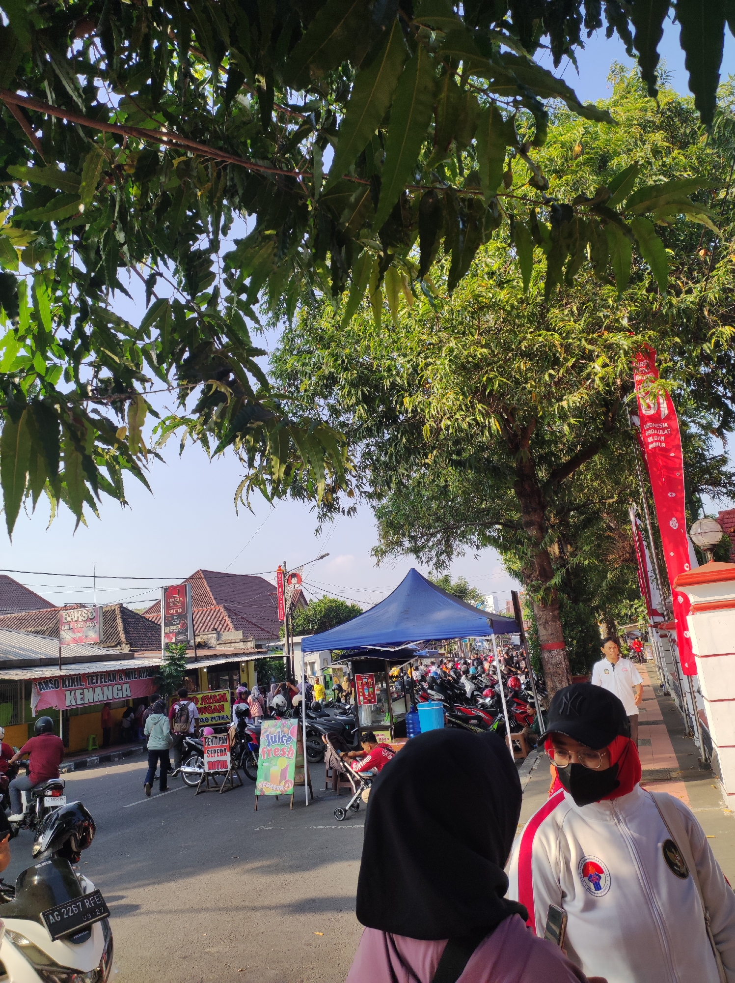 In the city of Blitar, Indonesia, an outdoor street scene unfolds under dappled sunlight filtering through leafy trees. The foreground features the backs of two individuals, one wearing a black hijab and a pink garment, and the other a white jacket with red trim, a black baseball cap, glasses, and a face mask. They appear to be in conversation. To their right, a paved sidewalk is bordered by a white wall with a red accent, and a white streetlight. Further back, a large tree stands prominently.
The street is lined with shops and stalls. A blue tent covers a vendor area, filled with parked motorcycles. Banners are visible, including a red one with Indonesian text and white graphics, and another advertising "BAKSO KIKIL, TETELAN & MIE AYAM KENANGA". Signs for "BAKSO" and "MAKAN AYAM" are also present. People are seen walking and gathered around the stalls. The ground is asphalt, and a white motorcycle with a license plate reading "AG 2267 RFE" is parked in the lower left corner. The sky is clear blue.