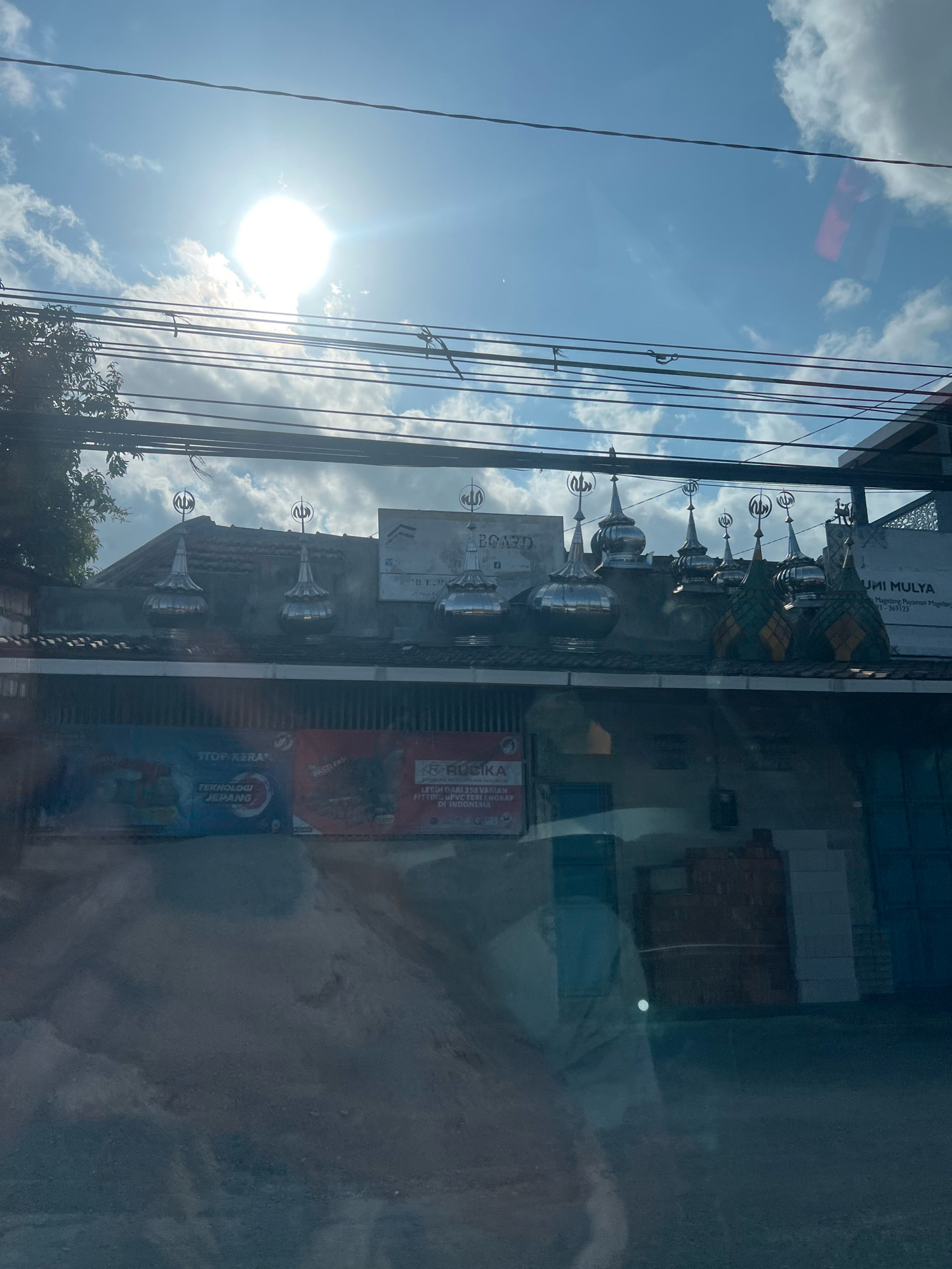 The photograph captures a street view in Payaman, Indonesia, dominated by a bright sun in a partly cloudy sky. Several utility wires crisscross the frame horizontally, with some appearing to sag. Below the wires, the roofline of a building is visible, adorned with multiple decorative finials resembling mosque domes. These finials are a mix of silver-toned and green and yellow patterned structures, topped with Arabic calligraphy. Two prominent signs are affixed to the building's facade. One sign, predominantly blue and red, reads "STOP KERAMIK TEKNOLOGI JEPANG" (Stop Ceramics Japanese Technology). Another sign, to the right, appears to be a business name, "UMI MULYA" followed by smaller text. The foreground is blurred, showing a reflective surface that appears to be the interior of a vehicle, possibly a car or bus, with a faint silhouette of a person visible. The overall impression is of a commercial or religious building situated along a street on a sunny day.