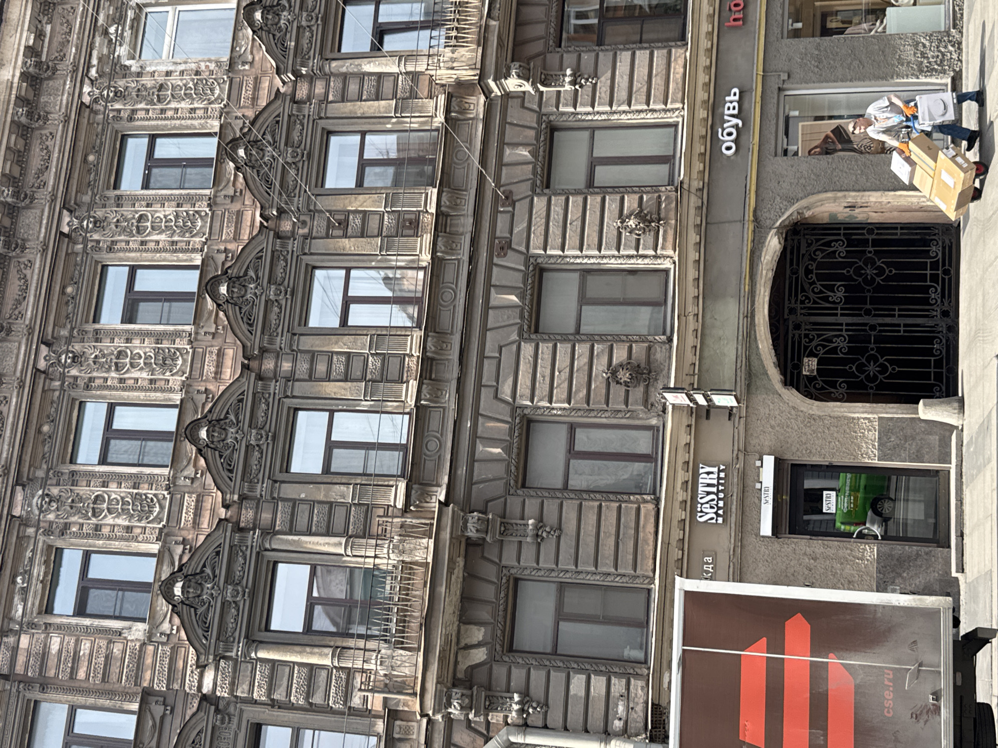 This image depicts an ornate building in Saint Petersburg, Russia. The building has multiple floors with numerous windows, each framed by intricate architectural details. The facade is primarily gray, suggesting stone or concrete construction. To the right of the building's center is an arched entrance with decorative ironwork gates. A person, possibly a delivery worker, is visible carrying boxes near the entrance. Commercial signage indicates the presence of shops, one labeled "обувь," which translates to "shoes" in Russian. Another sign reads "Sestry Mamutiny." The environment is urban, with typical city street features.