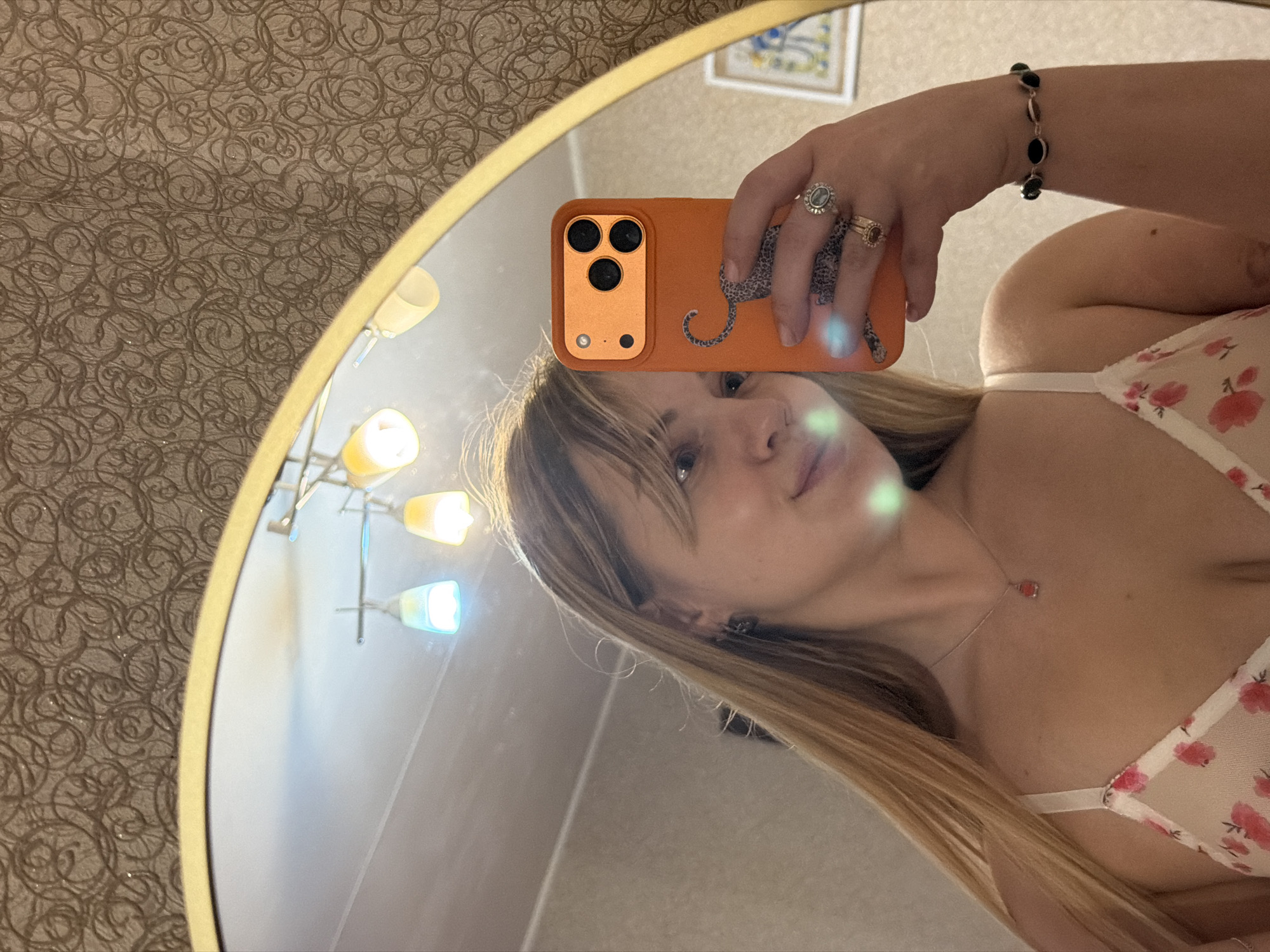 A woman is taking a selfie in a mirror. The mirror has a gold frame and reflects a room with patterned wallpaper and a chandelier. The woman is holding an orange phone with a leopard design on the case. She is wearing a floral top, a necklace, a bracelet, and rings. The location is unknown, but the city is Naberezhnye Chelny, Russia.