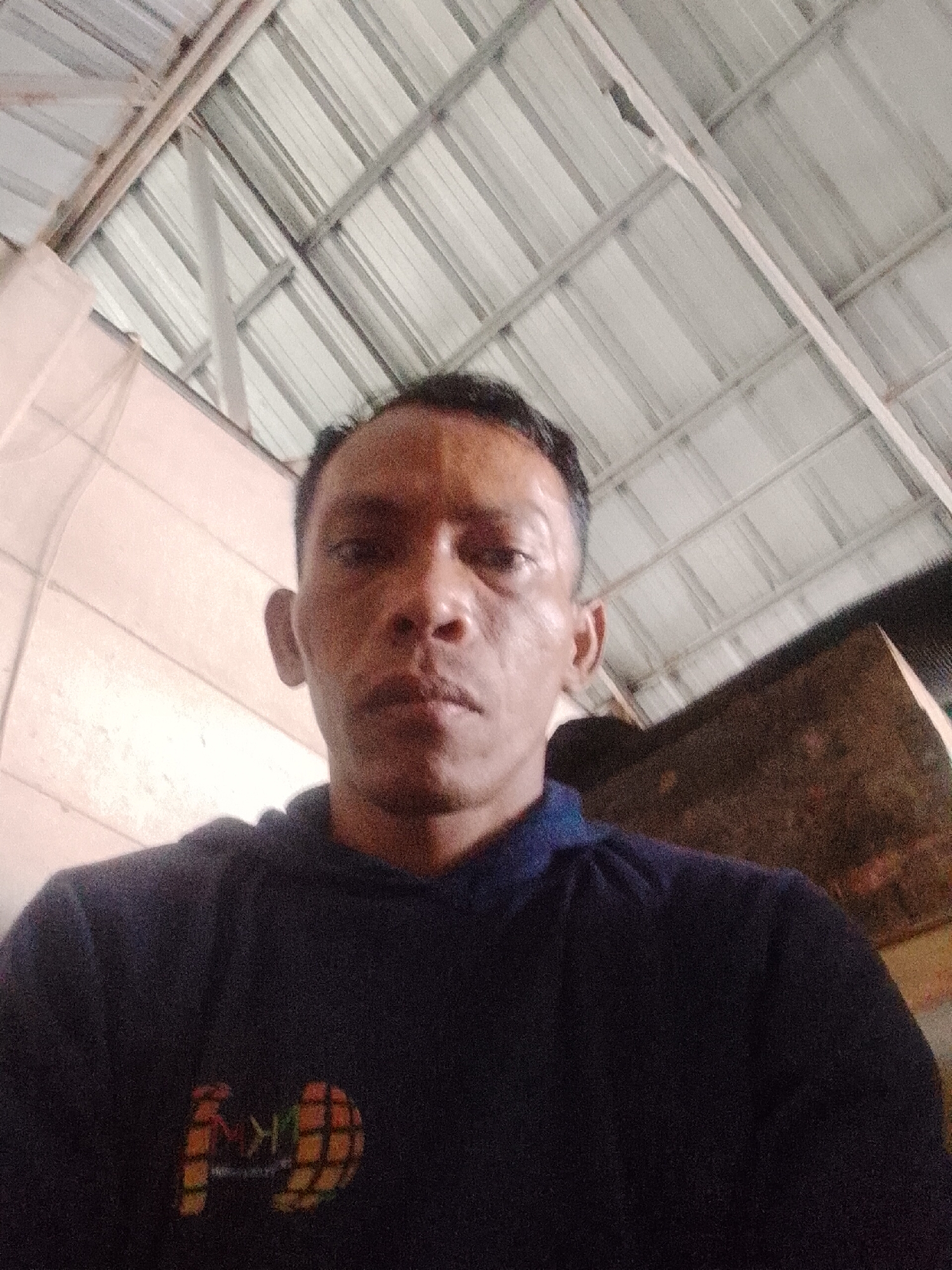 A medium shot depicts a man wearing a dark hooded sweatshirt, looking directly at the camera. The sweatshirt has a graphic on the lower left side. The background consists of an interior ceiling with metal supports and panels, typical of a simple roof structure. A partial wall is visible on the right. The location is Bukit Ketok, Indonesia.