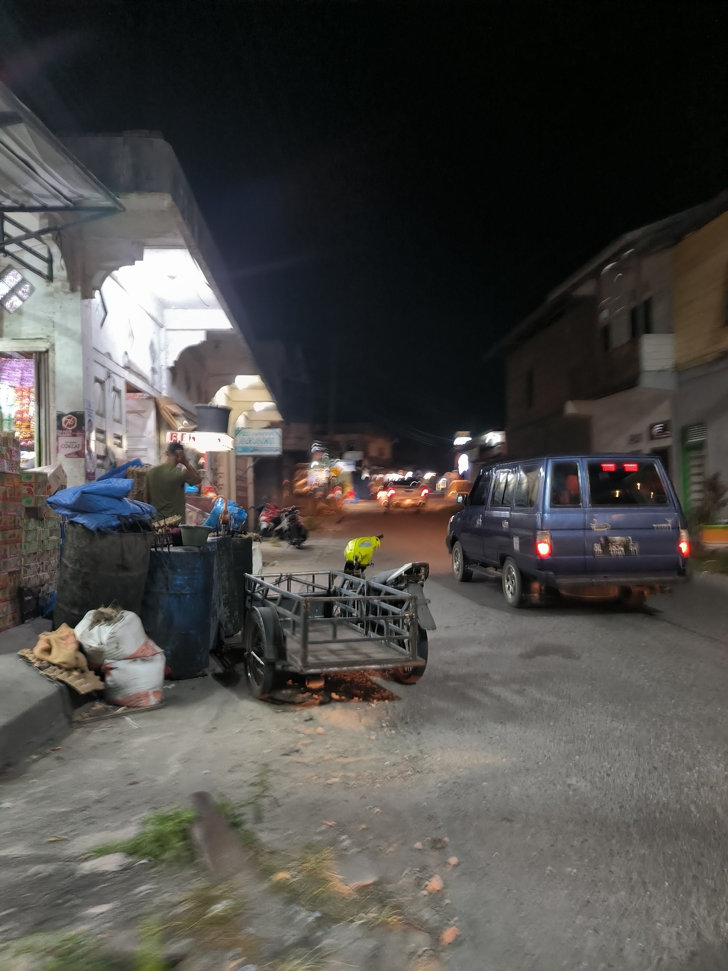 The image depicts a nighttime scene on a street in Simpang Tiga Redelong, Indonesia. In the foreground, to the left, are several large sacks, some appearing to be burlap, piled on the ground. Behind these sacks are two large, dark barrels, one partially covered with a blue tarp. A small, metal-framed cart with a cargo bed is positioned to the right of the barrels.
Further down the street, a blue minivan is visible, traveling away from the camera. Its brake lights are illuminated. The street itself is paved and appears to be an urban or semi-urban setting, with buildings lining the right side of the road. The overall lighting suggests it is night, with artificial light sources casting illumination. To the left, the facade of a building is visible, with some items displayed inside a storefront. Another person can be seen standing near the barrels on the left, facing away from the camera and talking on a phone. There are also a few motorcycles parked further down the street. The scene appears to be a street-side area with a mix of parked goods and moving vehicles.