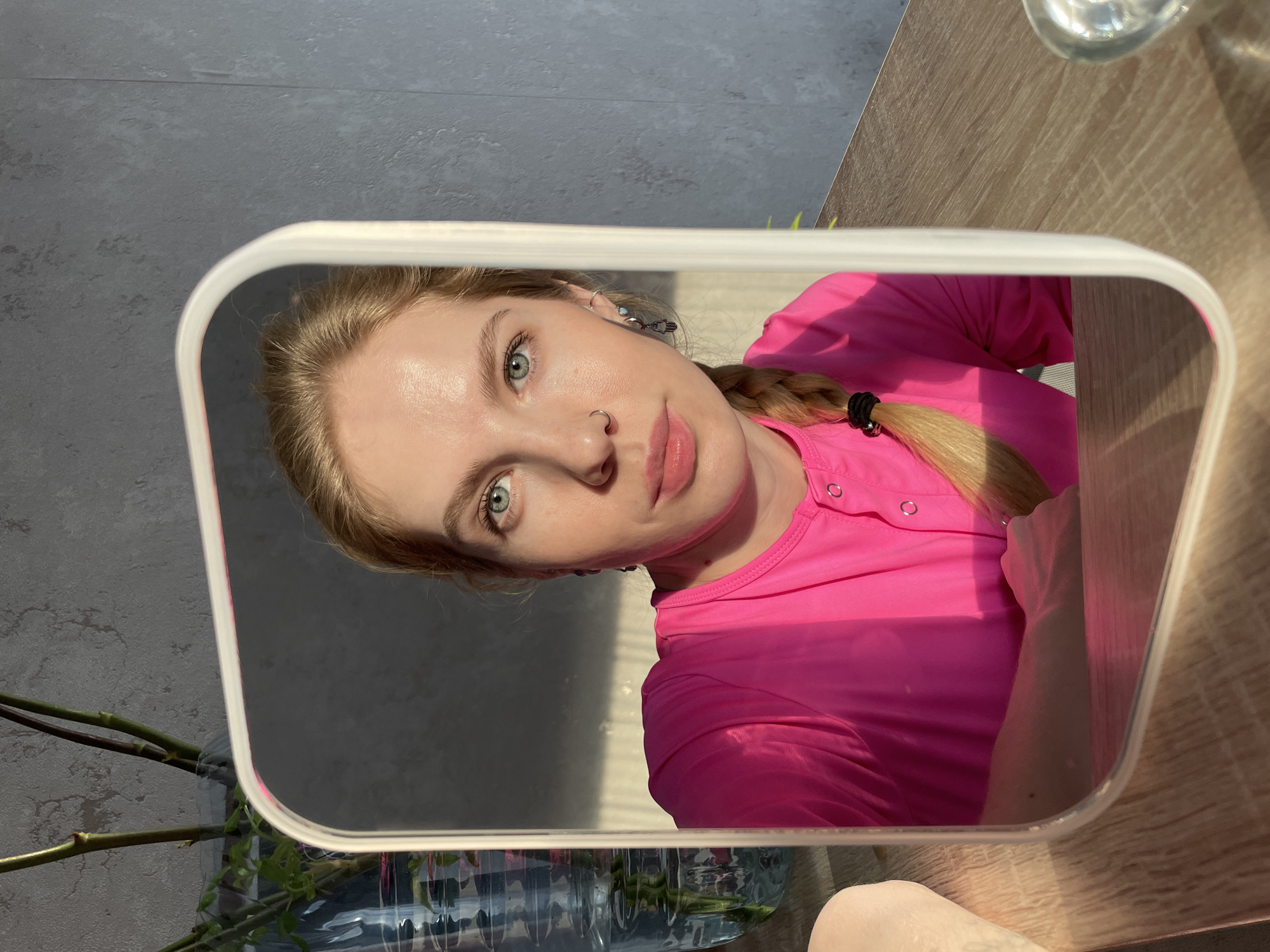 The image shows a fair-skinned woman with light blonde hair, styled in a braid, wearing a bright pink button-down shirt. She is looking at the camera through a mirror she is holding. The mirror's frame is white. Her facial features include green eyes, a nose ring, and multiple earrings. Behind the mirror, there is a vase with plants, set on a wooden table. The background consists of a gray wall and part of a desk. It is difficult to confirm that this image was taken in Izhevsk, Russia as there is no specific information to identify the city or location.