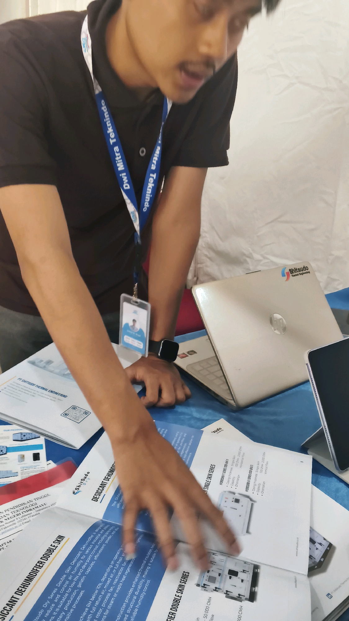 An overhead shot captures a man in a dark brown polo shirt with a lanyard displaying "Dwi Mitra Teknindo" and a name tag. He is leaning over a table covered with a blue cloth, his left hand resting on a brochure. In front of him, a silver laptop with the "Shitaudo Thermal Engineering" logo is open. To the right, a dark tablet is partially visible. Several brochures are spread across the table, one of which shows the title "DESICCANT DEHUMIDIFIER DOUBLE SKIN". The background is a plain white, slightly creased surface, likely a tent or display backdrop. The scene appears to be at an exhibition or trade show in Lohbener, Indonesia, given the text visible on the name tag and brochure.