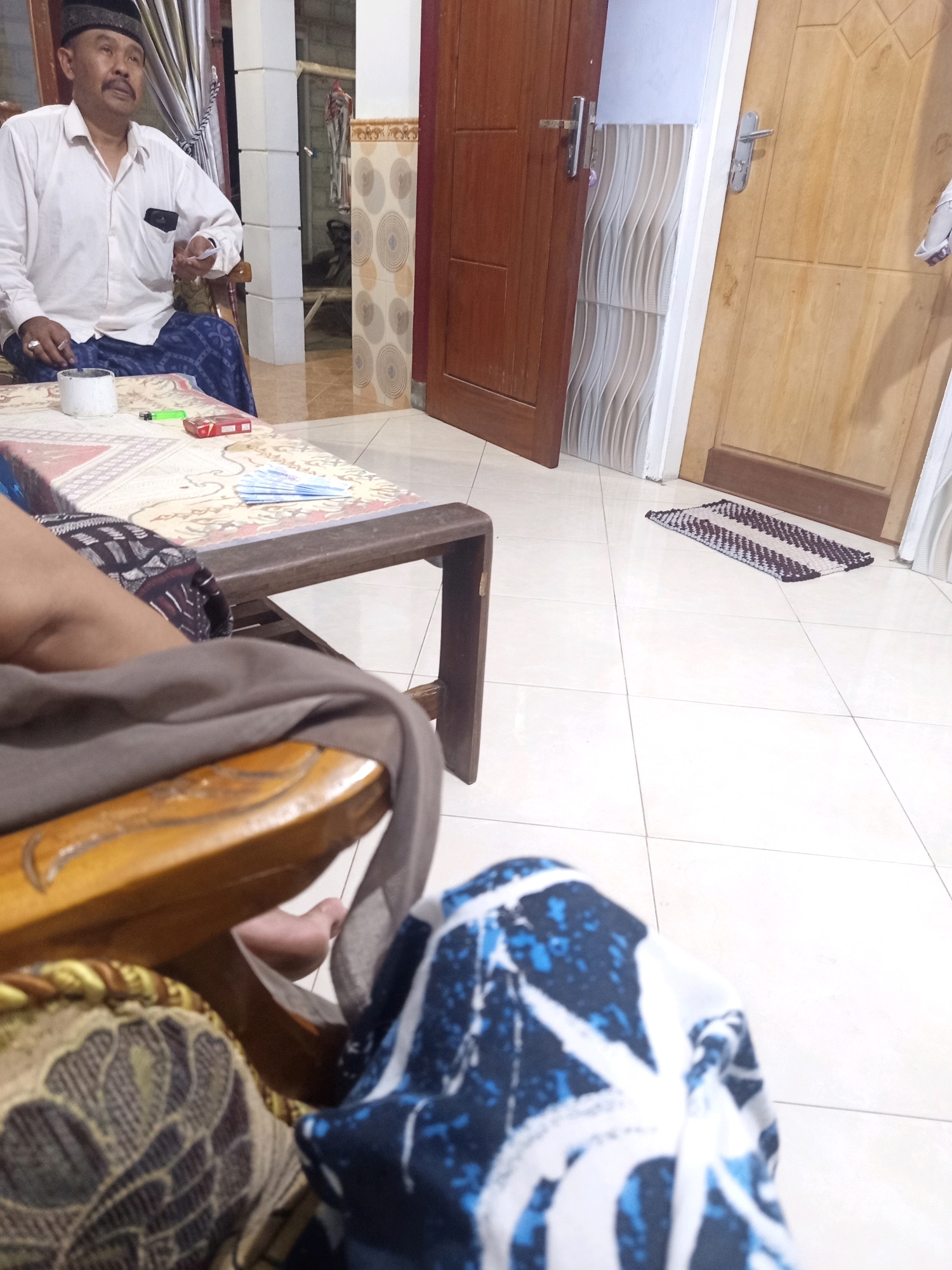 The image shows an indoor scene at night in Rowo Indah, Indonesia. In the foreground, a person is seated in a wooden chair, wearing blue and white patterned pants and a gray cloth draped over their lap. To the left, another person is seated, wearing a white button-down shirt, a black cap, and a blue patterned sarong. This person is holding a small white object and appears to be speaking. In front of them is a low wooden table covered with a patterned tablecloth. On the table, there is a white cylindrical object, a green lighter, and a red cigarette pack, along with several blue folded paper fans. The background reveals a wooden door and a lighter colored door, both ajar, revealing a hallway with tiled floors and patterned walls. A small striped rug lies on the tiled floor in front of the doors.