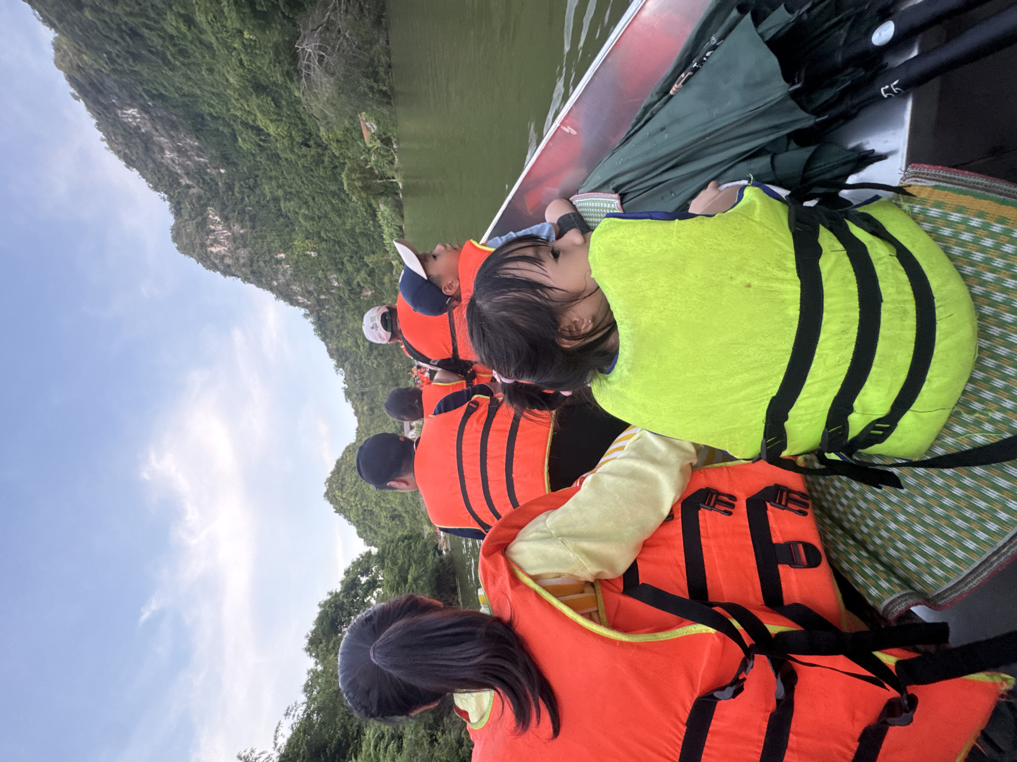 A boat filled with people, including children and adults, travels down a river with lush green mountains on either side. The people are wearing orange and green life vests for safety. The sky is cloudy with hints of blue. The boat appears to be in motion, as indicated by the water's reflection. This scene likely takes place in the karst landscape of Yên Sơn Ward, Vietnam, known for its scenic rivers and mountains.