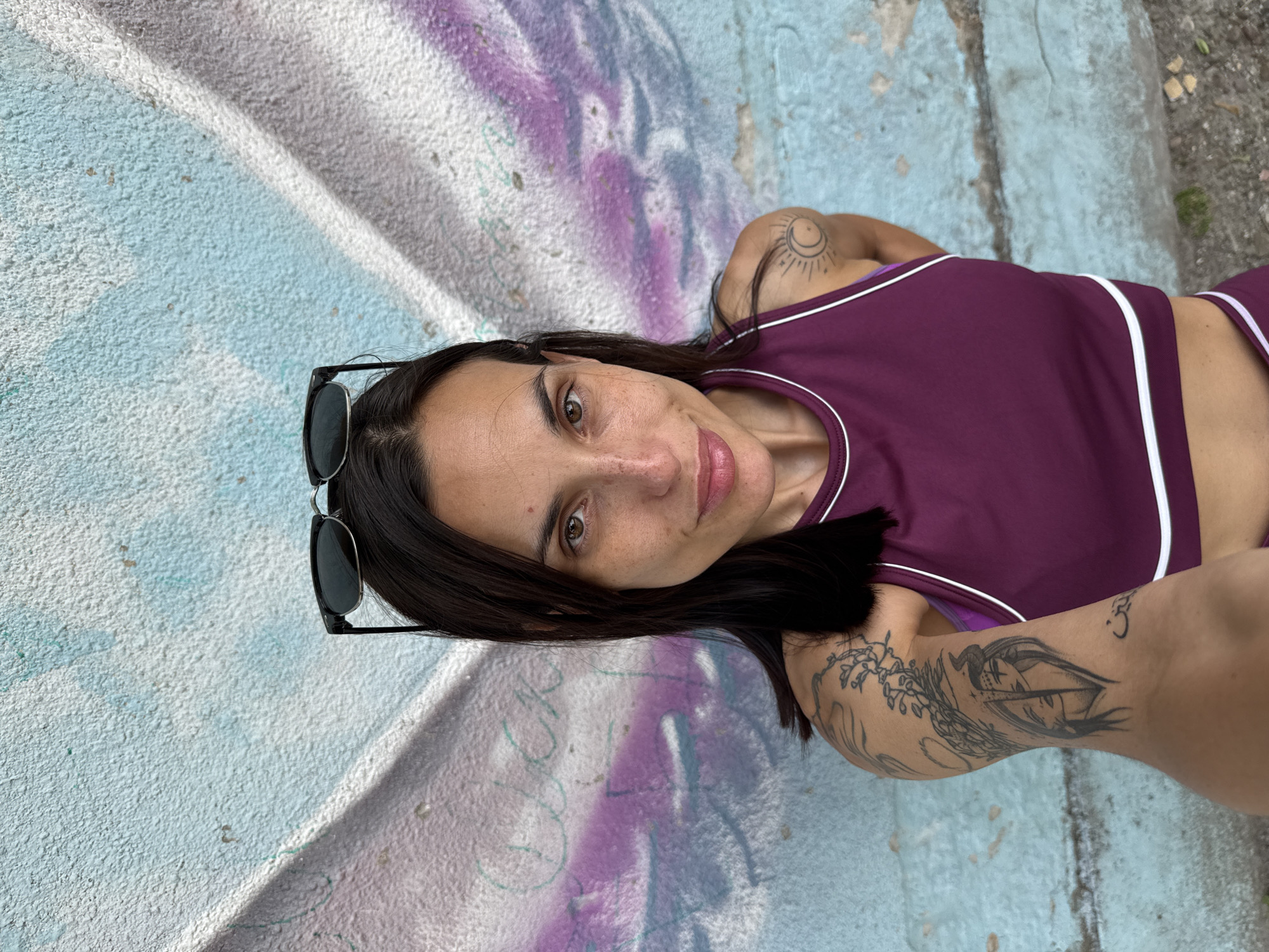 A woman with dark hair is positioned against a painted concrete wall. The wall features sections in light blue and purple with hints of green and white, and has a rough texture. The woman wears a dark purple athletic top with white trim and matching bottoms. Sunglasses are perched on her head. The woman has noticeable tattoos on her left arm and shoulder, depicting patterns and images. She is looking directly at the camera with a slight smile. The location appears to be outdoors, potentially an urban area where street art or graffiti is present. The overall impression is casual and informal, suggesting a candid snapshot. It is not possible to ascertain from the image whether the scene is located in Pleven, Bulgaria.