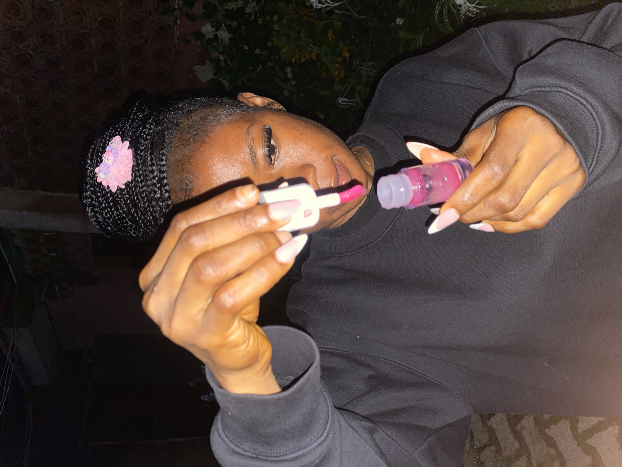 A medium shot captures a young Black woman in the foreground, holding a small bottle of lip gloss. She is looking down at the bottle, her lips slightly parted, as she applies the gloss with the wand. Her braided hair is pulled up into a bun, adorned with a pink flower clip. She is wearing a black sweatshirt with a crew neck. Her fingernails are manicured in a pale pink and white French tip style. The background is dark and out of focus, suggesting it is nighttime. There are some blurred hints of greenery behind her. The overall impression is a casual, candid moment captured outdoors. The image is not set in Jos, Nigeria.