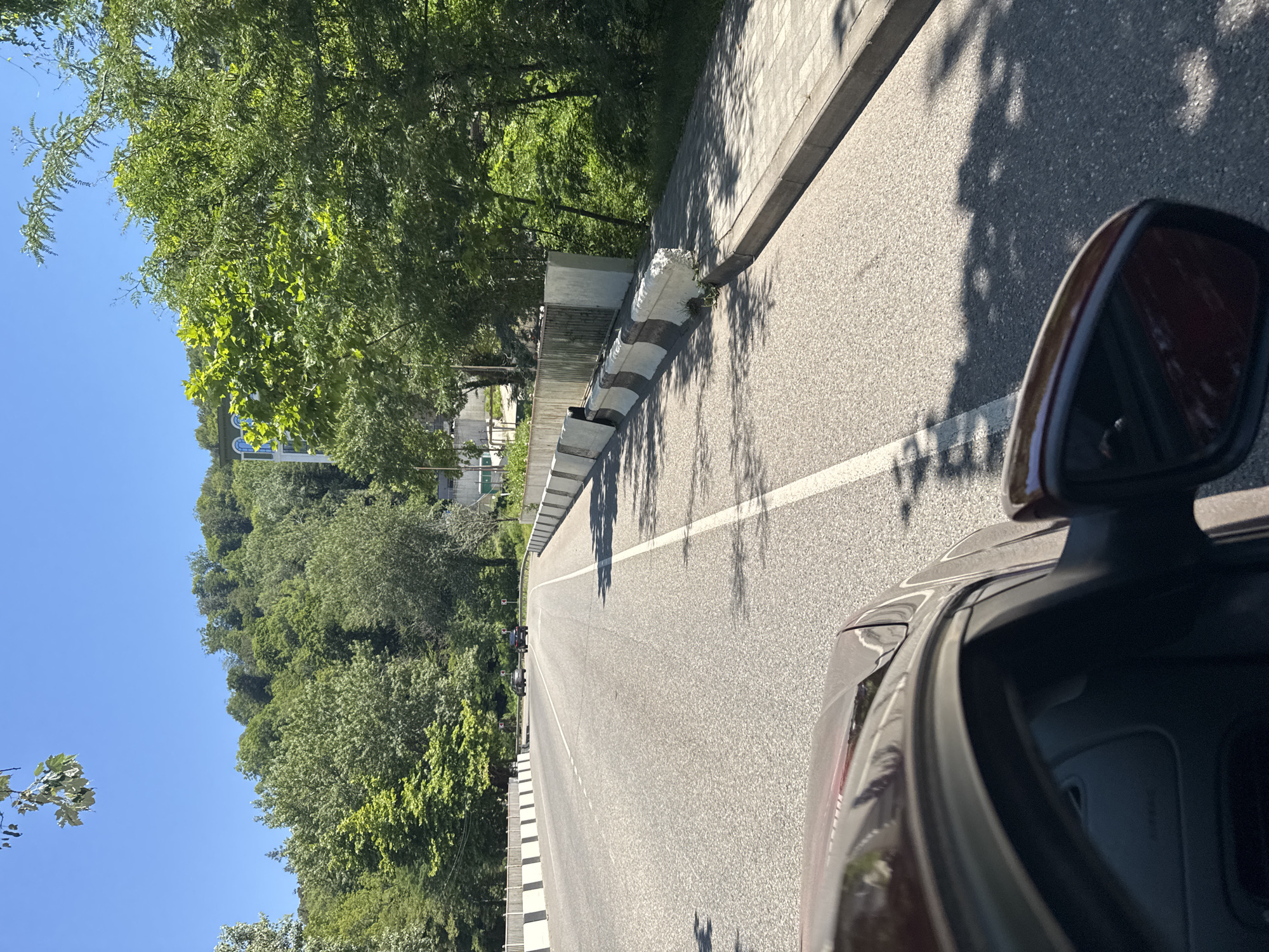 The image depicts a road in Sochi, Russia, on a sunny day. A dark-colored car is in the foreground, with its driver's side visible. The road ahead curves to the left and is bordered by a concrete barrier on both sides. Beyond the barriers, there are lush green trees and a hill. Two cars are visible in the distance on the road. The sky is clear and blue. The scene appears to be a scenic route with natural surroundings.