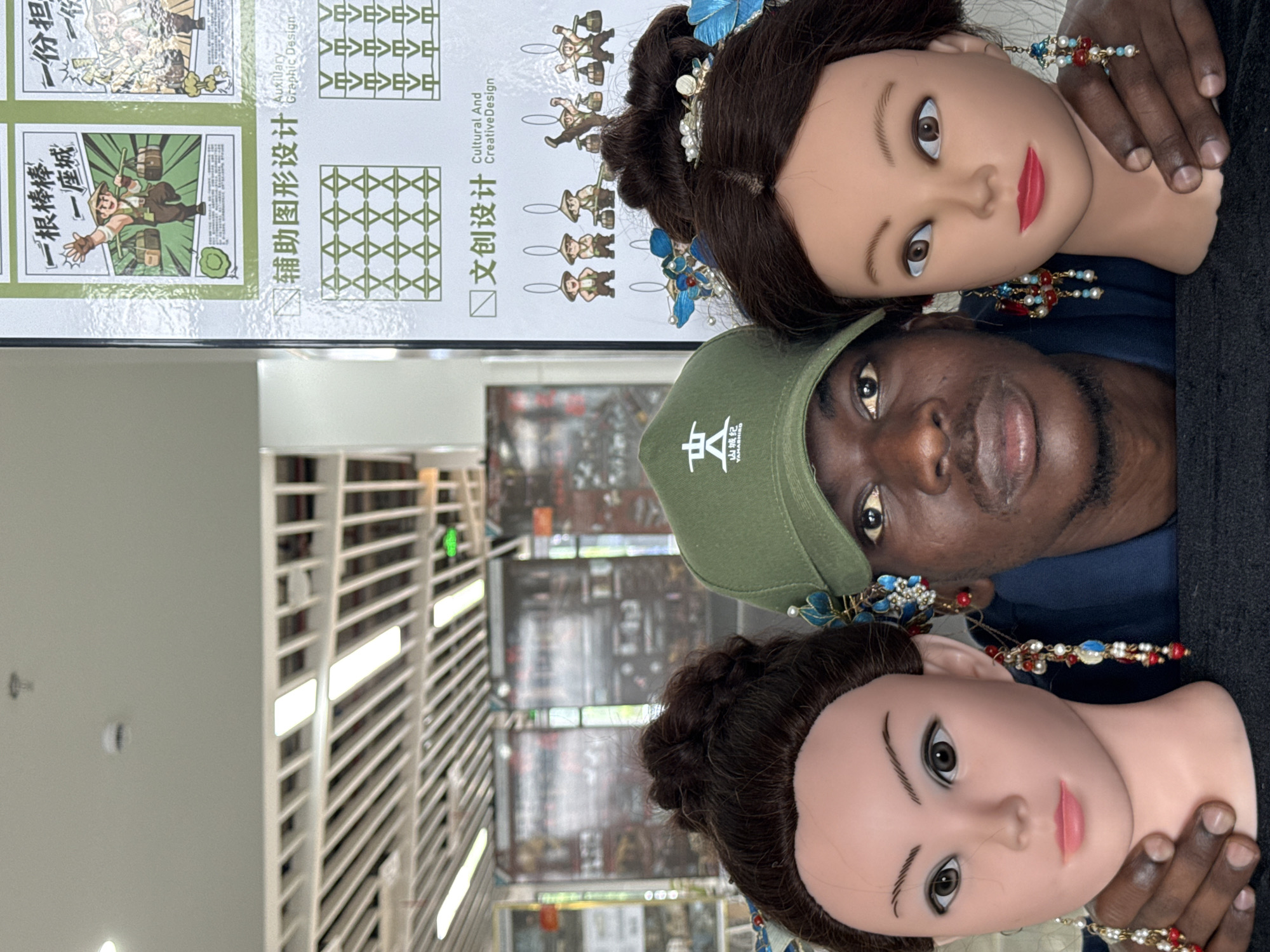 A Black man wearing a green baseball cap with a white logo stands between two female mannequin heads in what appears to be a retail or exhibition space in Jiangjin, China. The man's arms are around the mannequin heads, with his left arm supporting the left mannequin and his right arm around the right mannequin. The mannequins have dark brown hair styled in buns and are adorned with intricate, multi-colored earrings featuring blue feathers, pearls, and red beads. The man's eyes are looking directly at the camera, and his lips are slightly parted. Behind the man and mannequins, a large poster or display board is visible, featuring Chinese text and graphic designs, including illustrations of people carrying loads and geometric patterns. The environment suggests a commercial or cultural establishment.
