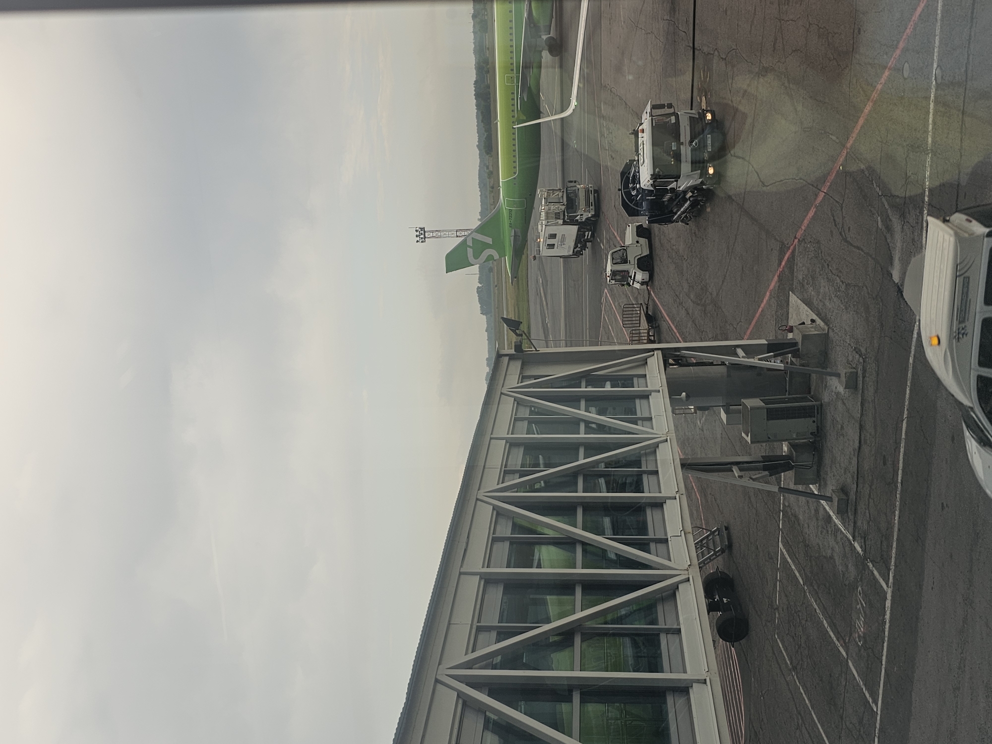 This is an outdoor, eye-level medium shot of an airport tarmac with a jet bridge in the foreground. The jet bridge is gray with large windows. Beyond the jet bridge, a bright green airplane with an "S7" logo on its tail is parked on the tarmac. Several service vehicles are present: a white truck with a black tank, a smaller white utility vehicle, and a white cargo van. The sky is overcast and hazy. The scene appears to be at Tolmachevo Airport (OVB) in Ob, Russia, based on the S7 Airlines livery and the general appearance of the airport infrastructure. There are no people clearly visible in the image. The tarmac shows cracks and markings.
