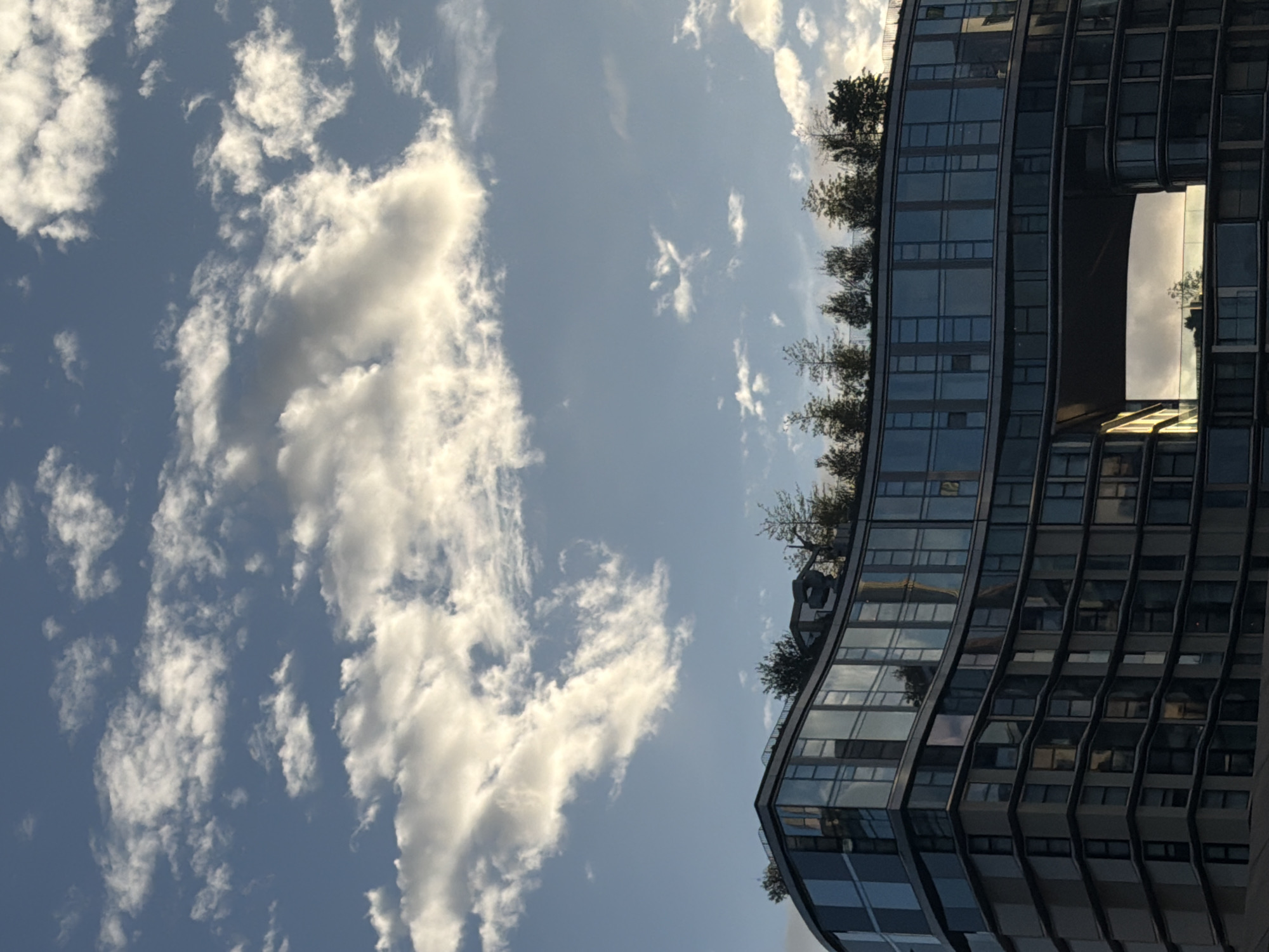 The image shows a tall, modern building with a curved facade and multiple windows, adjacent to a sky filled with white clouds. The building appears to have a large opening or cutout in the middle section, allowing the sky to be seen through it. The building also has a rooftop garden with trees and greenery. The building is in London, United Kingdom.